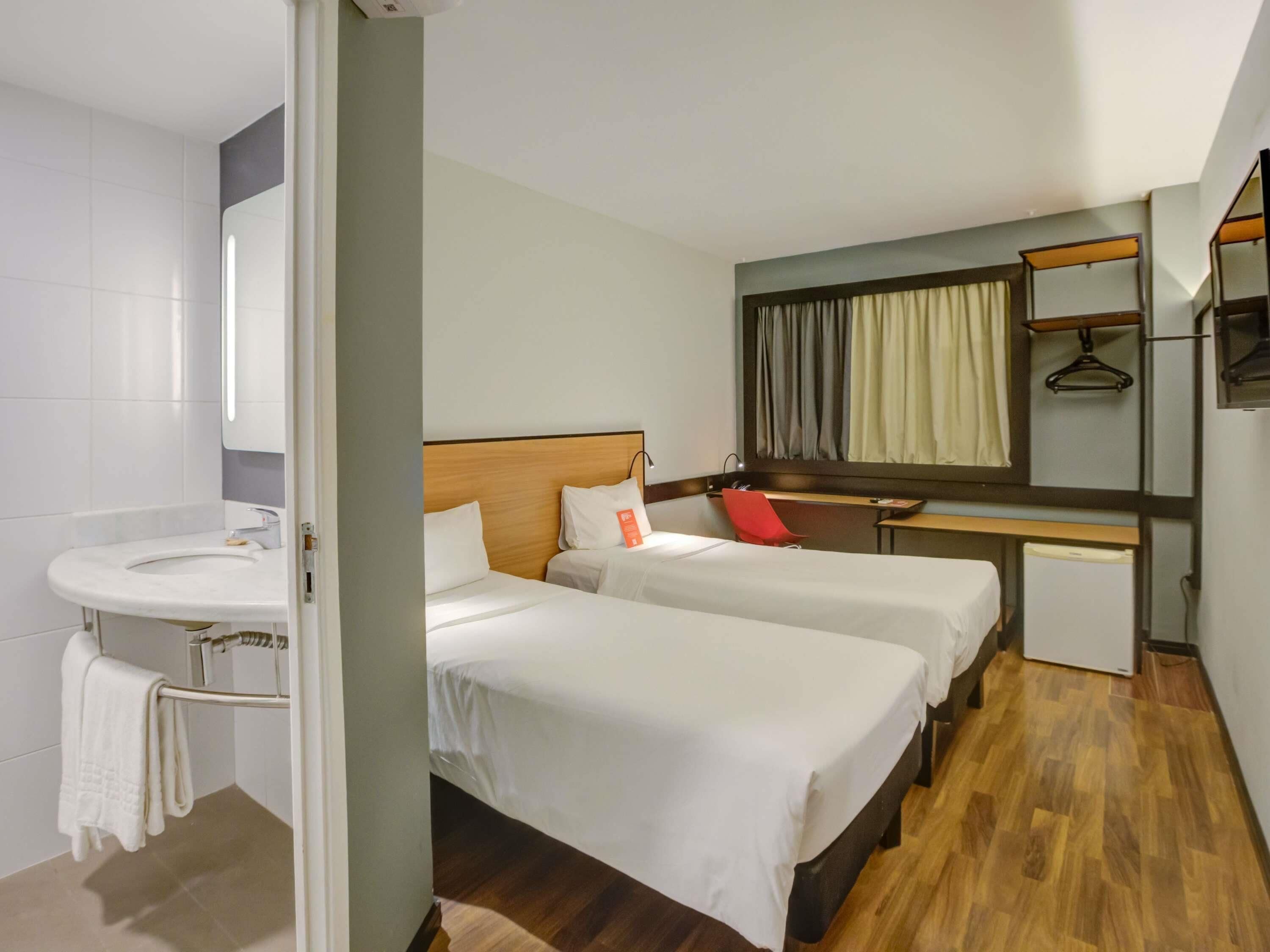 Ein Hotelzimmer mit zwei Betten, einem kleinen Schreibtisch, einem Spiegel und einem Badezimmer mit Waschbecken und Handtuchhalter.