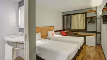 Ein Hotelzimmer mit zwei Betten, einem kleinen Schreibtisch, einem Spiegel und einem Badezimmer mit Waschbecken und Handtuchhalter.