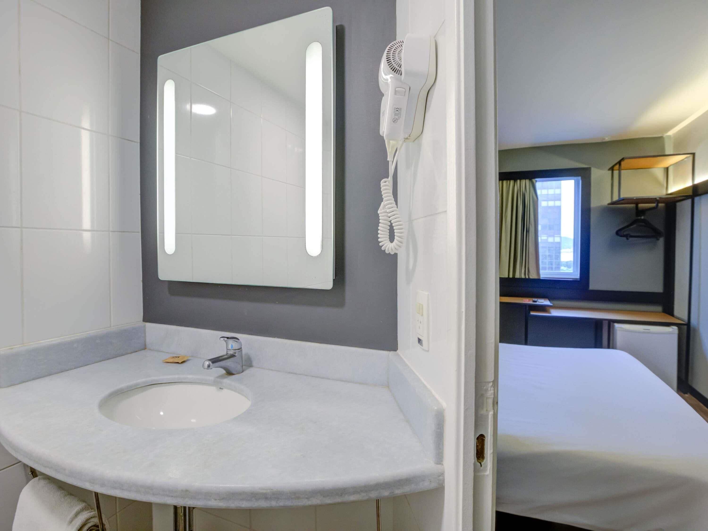 Une salle de bain moderne avec un lavabo blanc, un miroir doté de lumières intégrées et un sèche-cheveux fixé au mur.