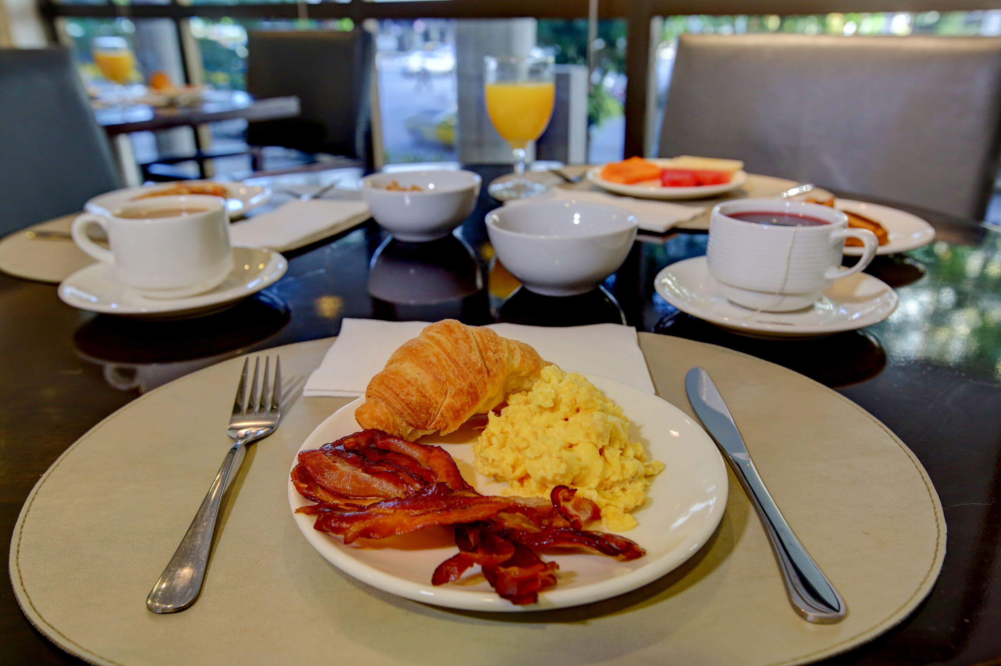 A breakfast setting with croissants, scrambled eggs, and bacon on a plate, accompanied by coffee and juice.