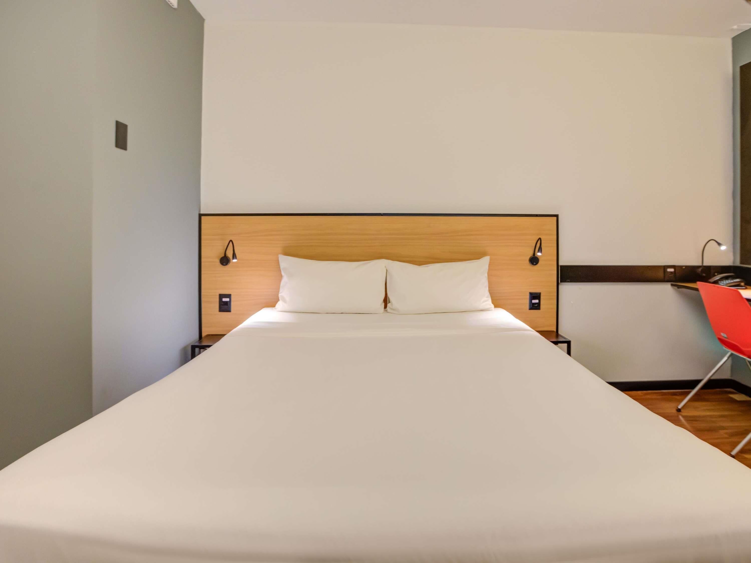 A neatly made bed with a wooden headboard, two bedside lamps, and a red chair to the right.
