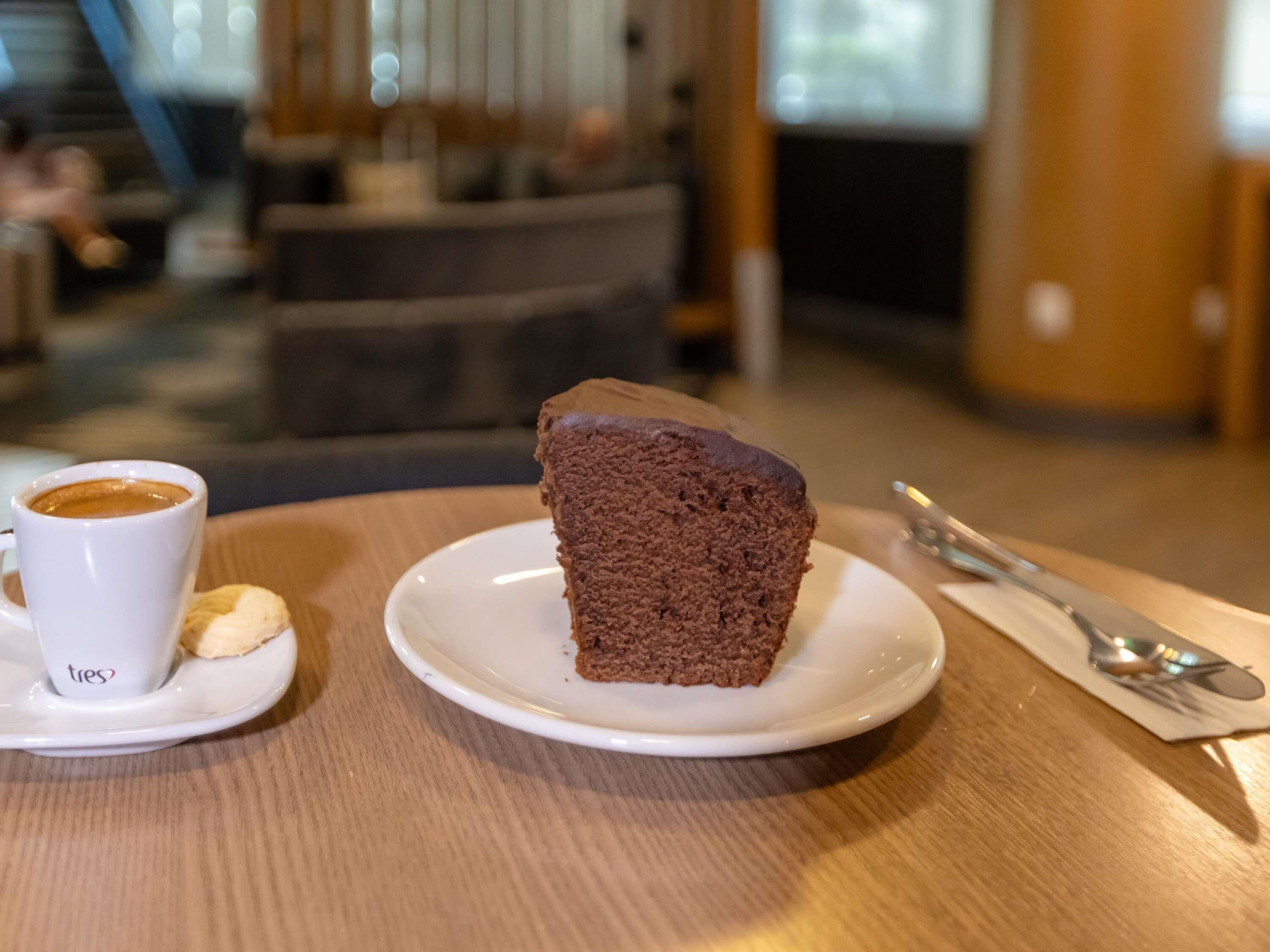A slice of chocolate cake on a white plate with a cup of coffee and a spoon on a wooden table.