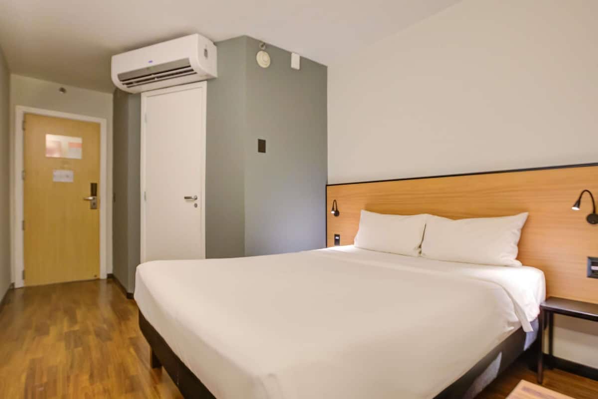 A hotel room with a large bed, a wooden headboard, a wall-mounted air conditioner, and a door with a sign.