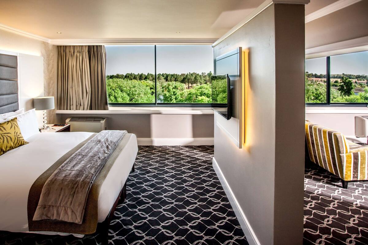 A hotel room with a large bed, a window with a view of greenery, a bathtub, and a TV mounted on the wall.