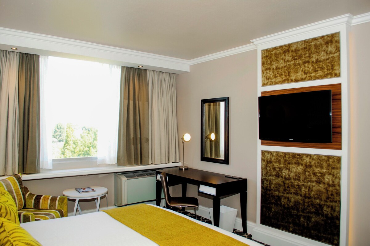 A hotel room with a bed, a desk, a chair, a small table, a television, and a window with curtains.