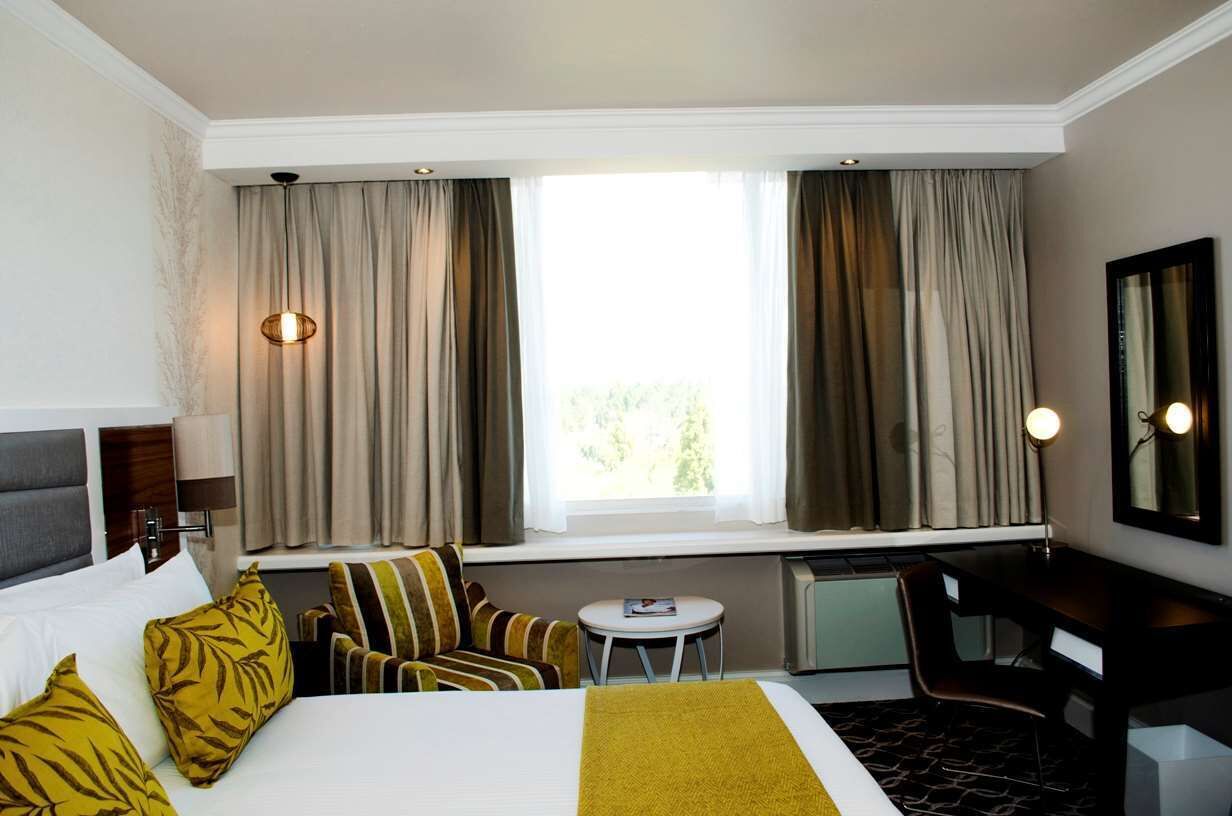 A hotel room with a bed, a desk, a chair, a small table, and a window with curtains.