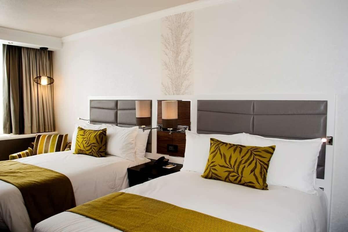 A hotel room with two beds, each with a grey headboard, white bedding, and a green throw blanket. There are two bedside tables with lamps, a wall-mounted TV, and a large window with curtains.