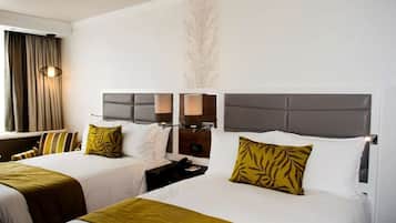 A hotel room with two beds, each with a grey headboard, white bedding, and a green throw blanket. There are two bedside tables with lamps, a wall-mounted TV, and a large window with curtains.