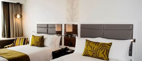 A hotel room with two beds, each with a grey headboard, white bedding, and a green throw blanket. There are two bedside tables with lamps, a wall-mounted TV, and a large window with curtains.
