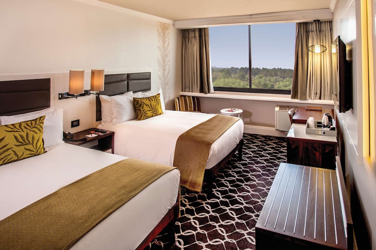 A hotel room with two beds, a desk, a chair, a TV, and a window with a view of trees.