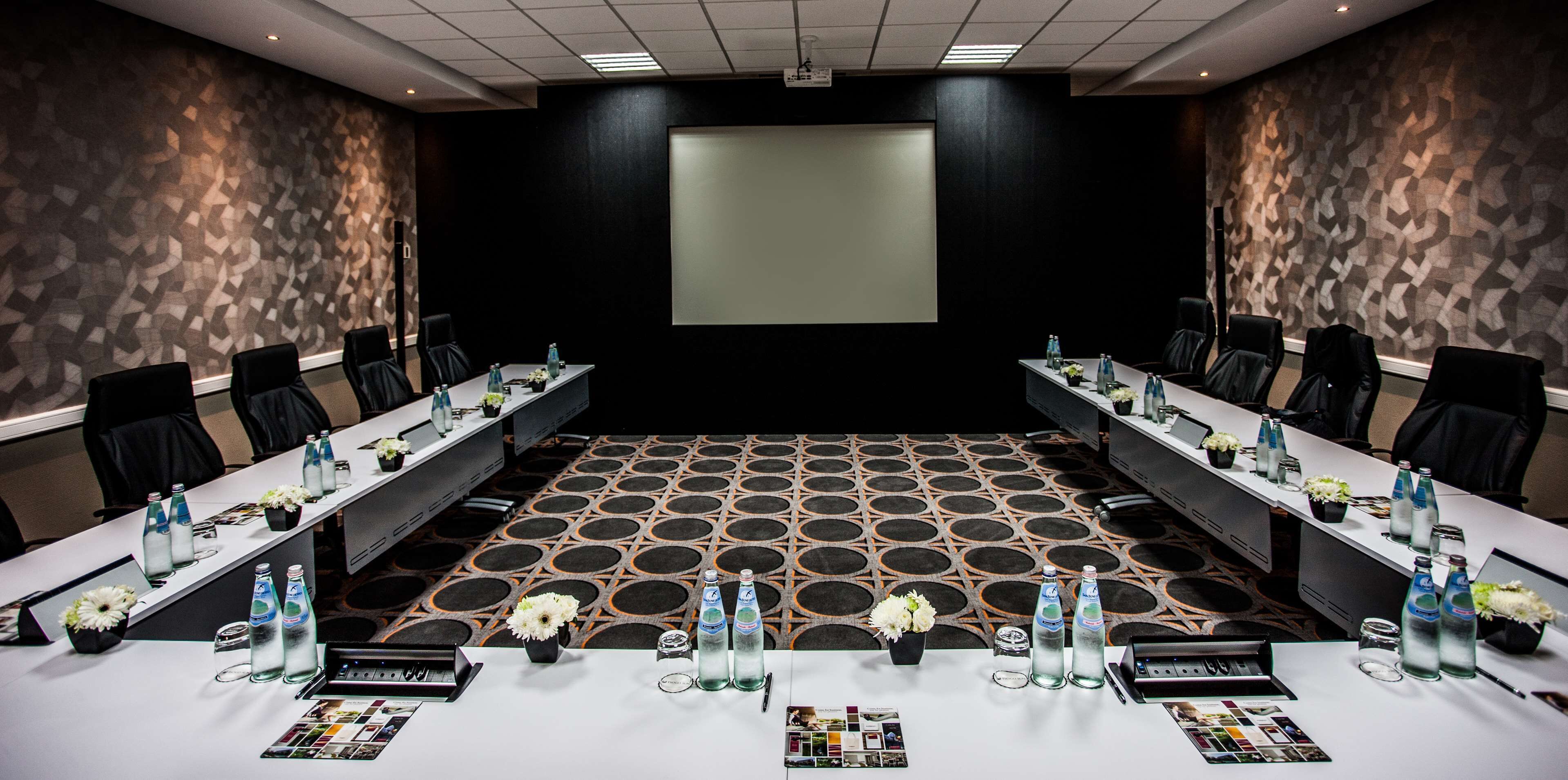 Un salón de conferencias con una gran mesa, sillas, botellas de agua y arreglos florales.