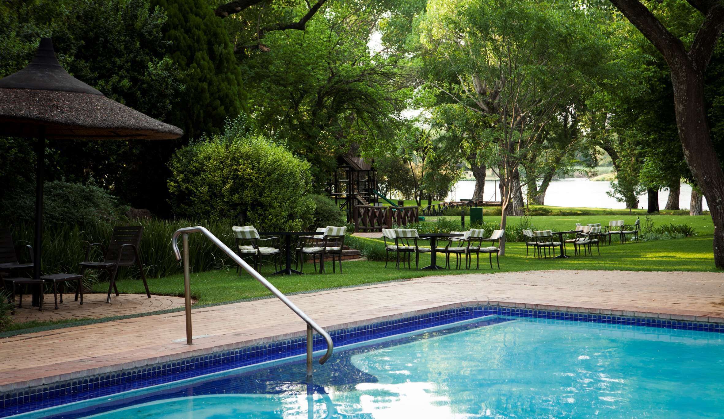A swimming pool with a tiled edge, surrounded by a patio area with chairs and tables, and a playground structure in the background.