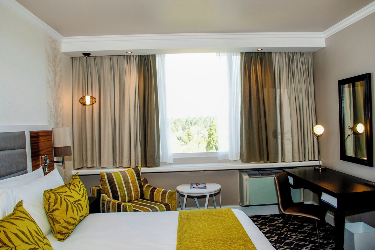 A hotel room with a bed, a desk, a chair, a small table, and a window with curtains.