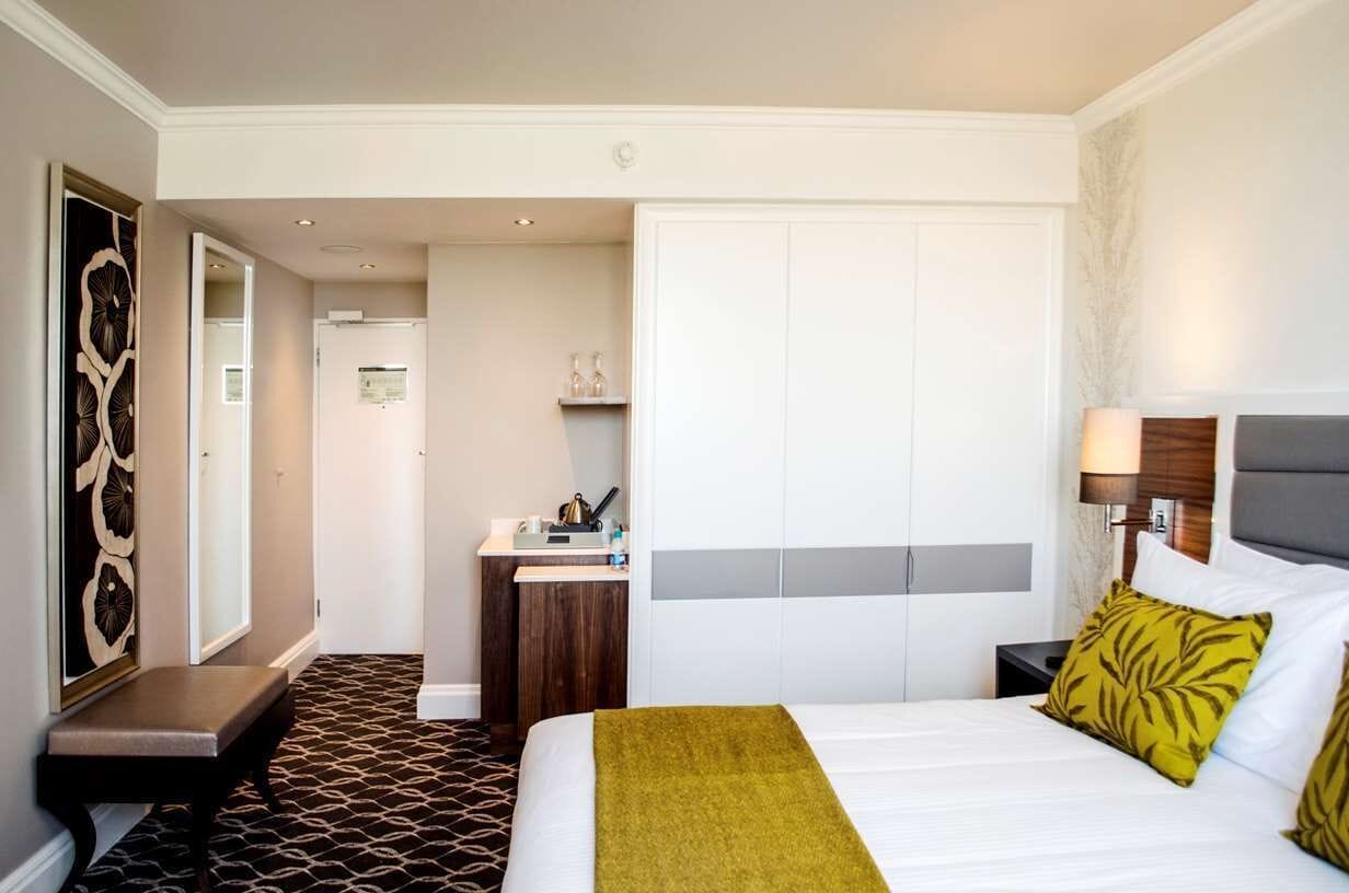 A hotel room with a bed, bedside table, lamp, and a small desk with a chair.
