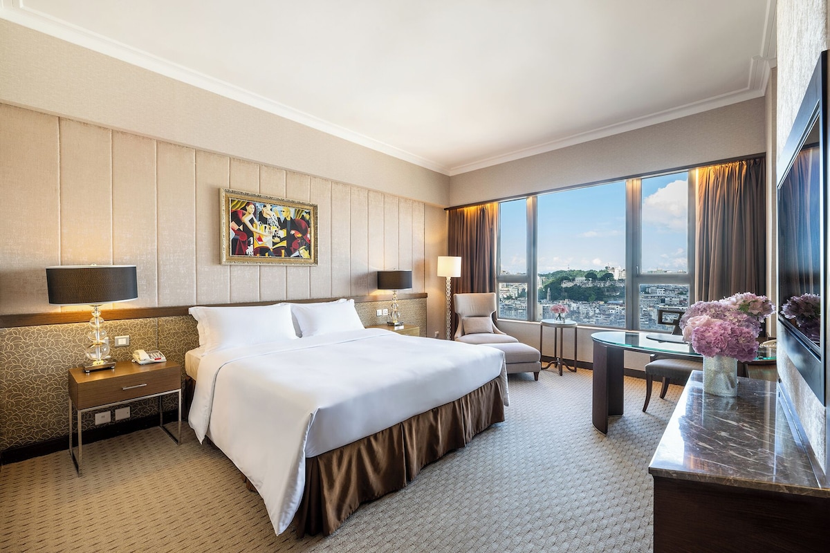 A hotel room with a large bed, a desk, a chair, a painting on the wall, and a view of the city.