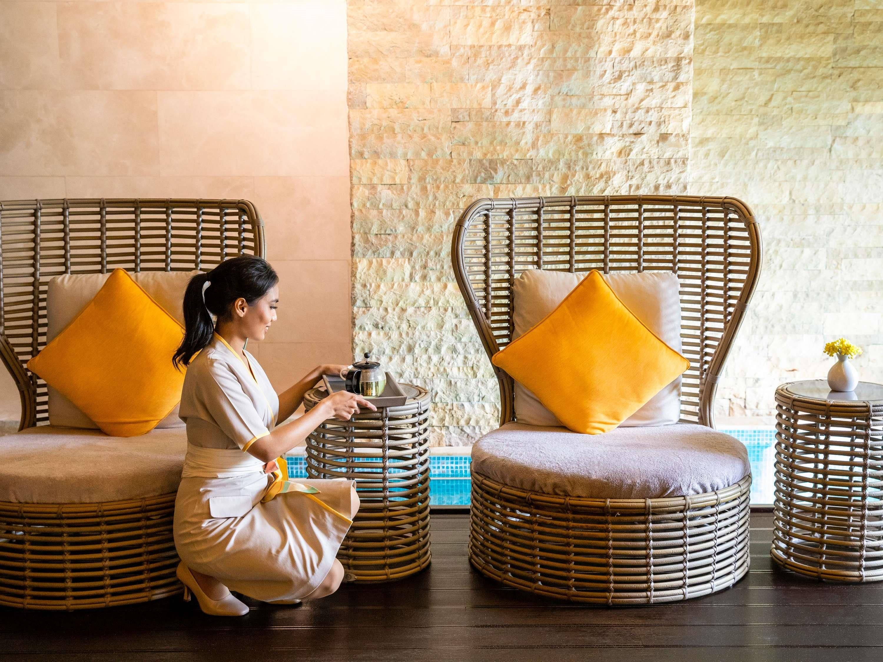 Une personne en uniforme, à genoux, se tient à côté d’une chaise en osier avec un coussin, d’un panier en osier et d’une petite table sur laquelle se trouve un vase.