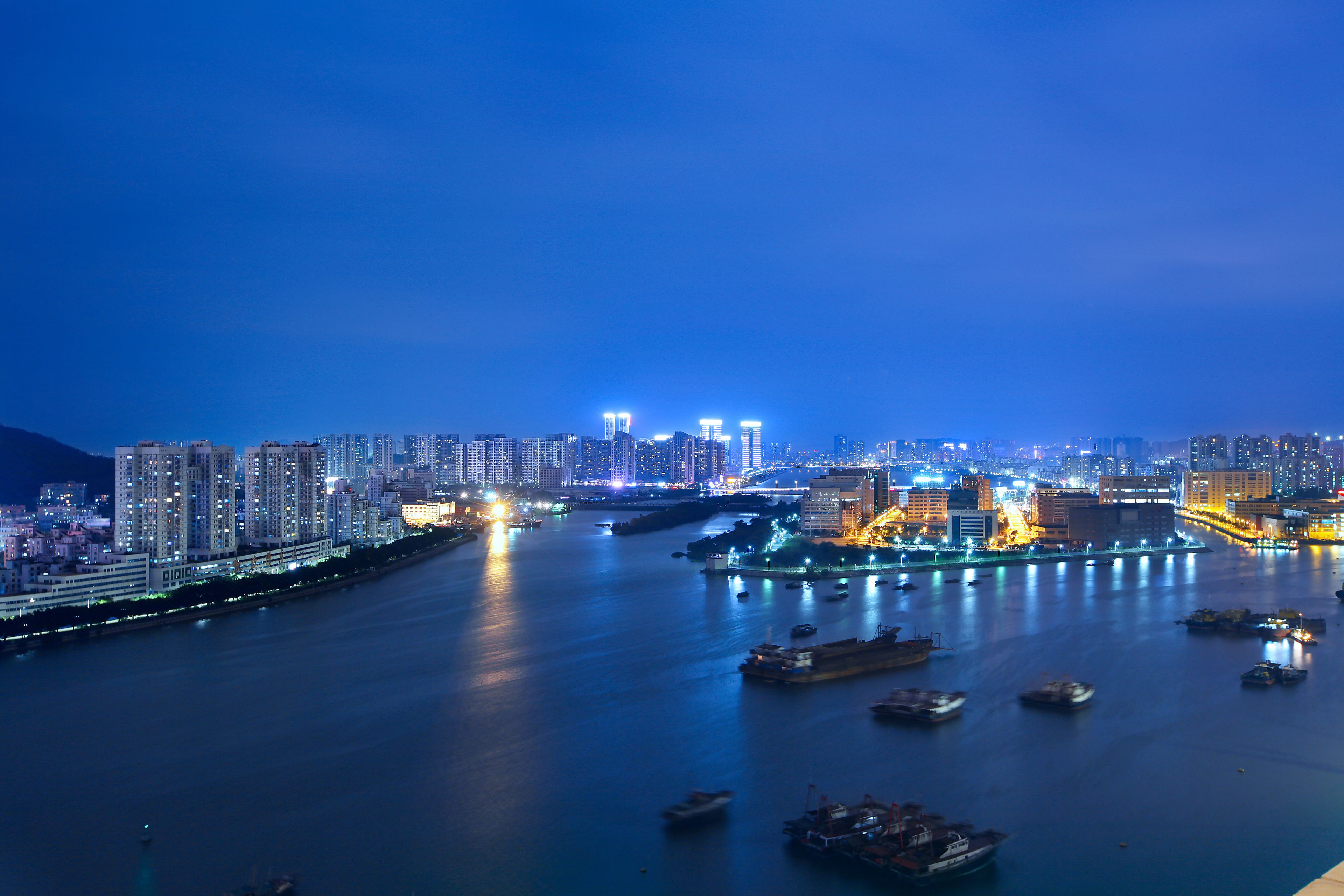 A nighttime cityscape with a river, illuminated buildings, and boats.