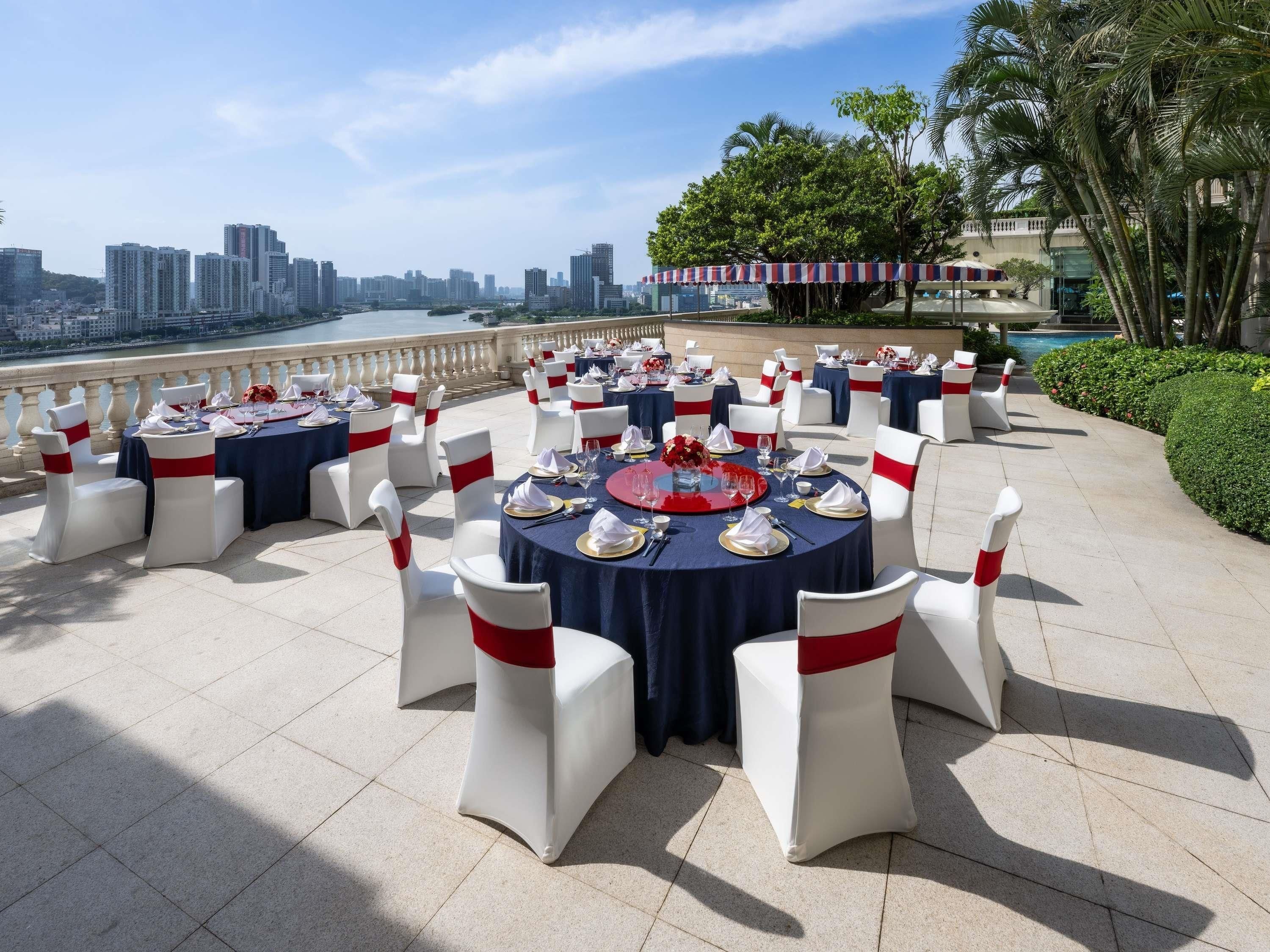 Montaje formal para comedor al aire libre con mesas redondas, sillas blancas y fajones rojos, con vista a un paisaje urbano y una masa de agua.