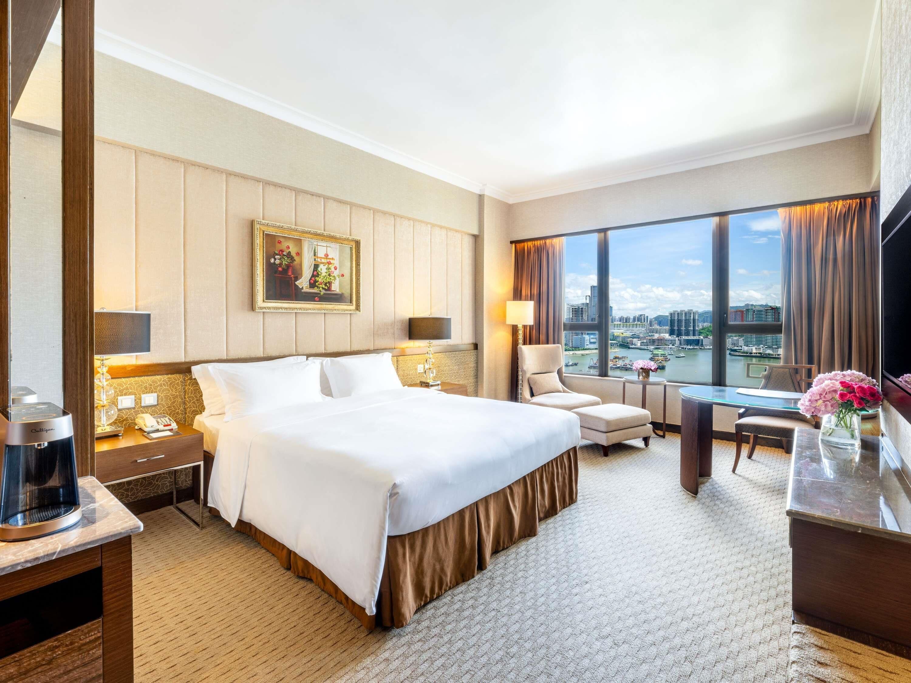 A hotel room with a large bed, a desk, a chair, a TV, and a view of the city.