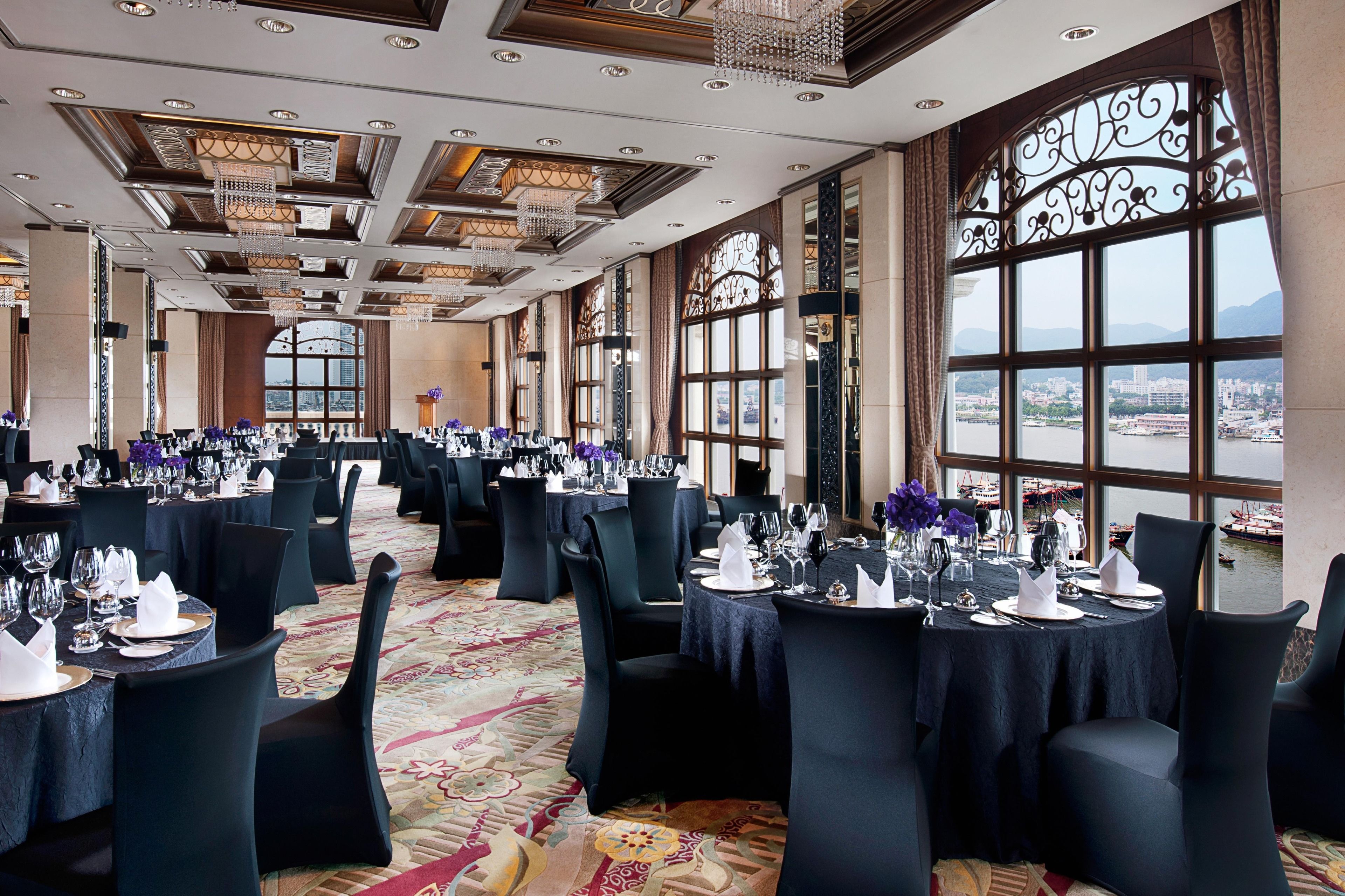 A banquet hall with round tables set for dining, large windows with decorative ironwork, and a view of a river and cityscape.