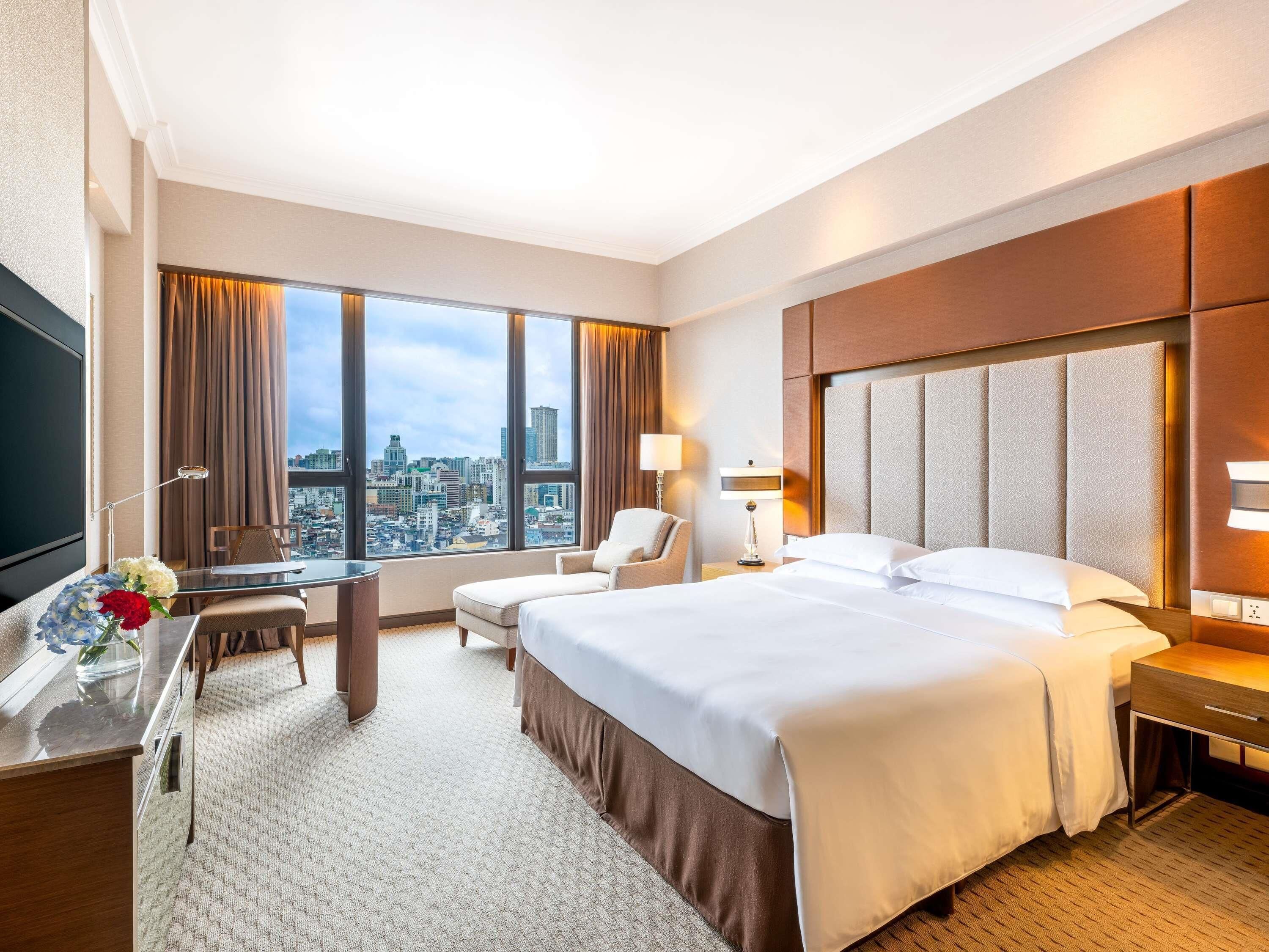 A hotel room with a large bed, a desk with a chair, a TV, and a city view.