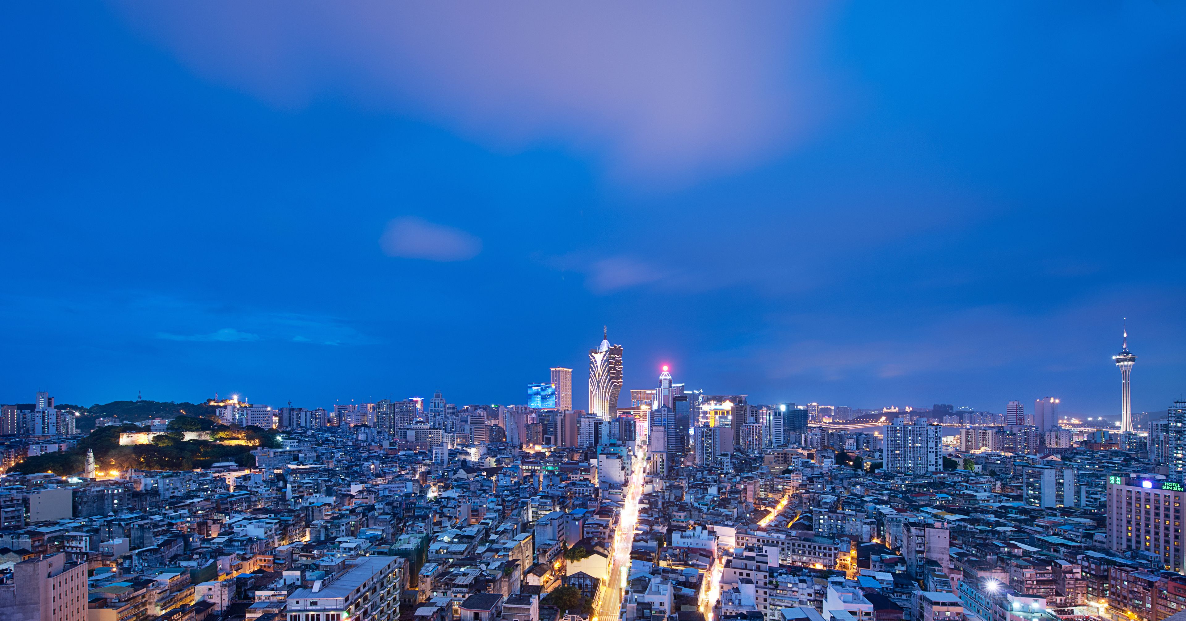 Un paisaje urbano al atardecer con imponentes rascacielos, una torre con un campanario característico y una torre con cables de suspensión.