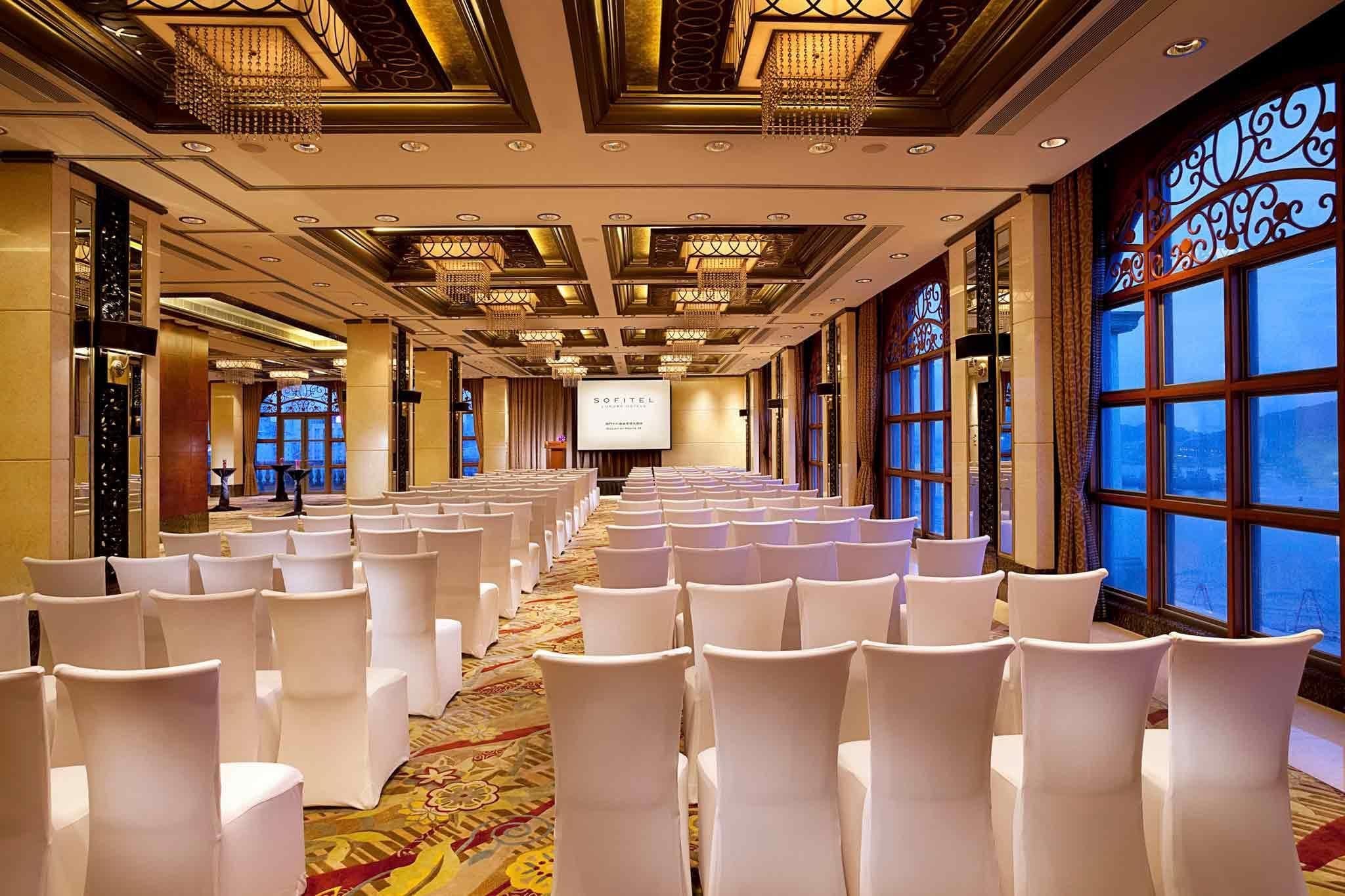 Un salón de conferencias con filas de sillas blancas, piso alfombrado, candelabros y ventanas amplias con diseños decorativos.