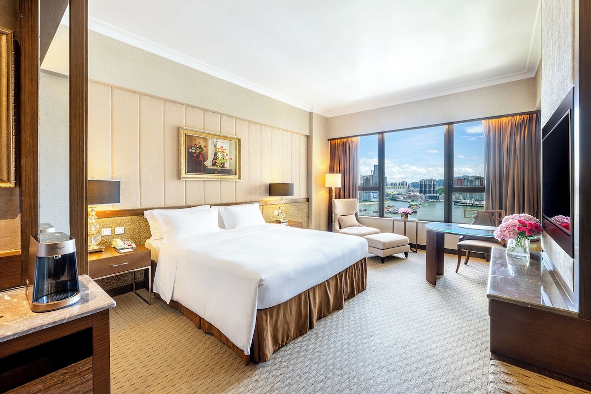 A hotel room with a large bed, a desk, a chair, a TV, and a view of the city.