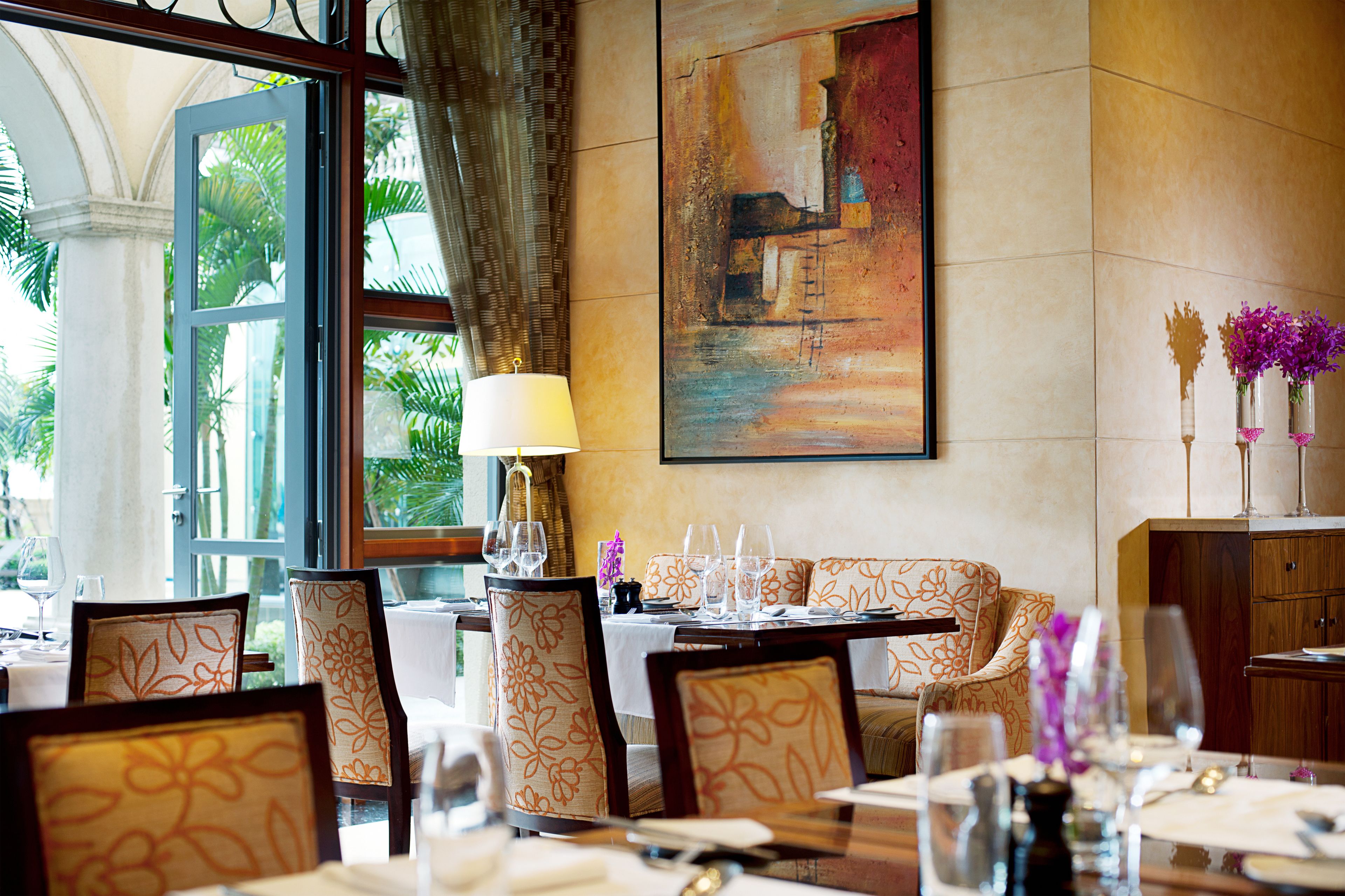 Comedor con una mesa dispuesta para una comida, sillas con tapicería floral, un cuadro en la pared y vistas a zonas verdes al exterior.