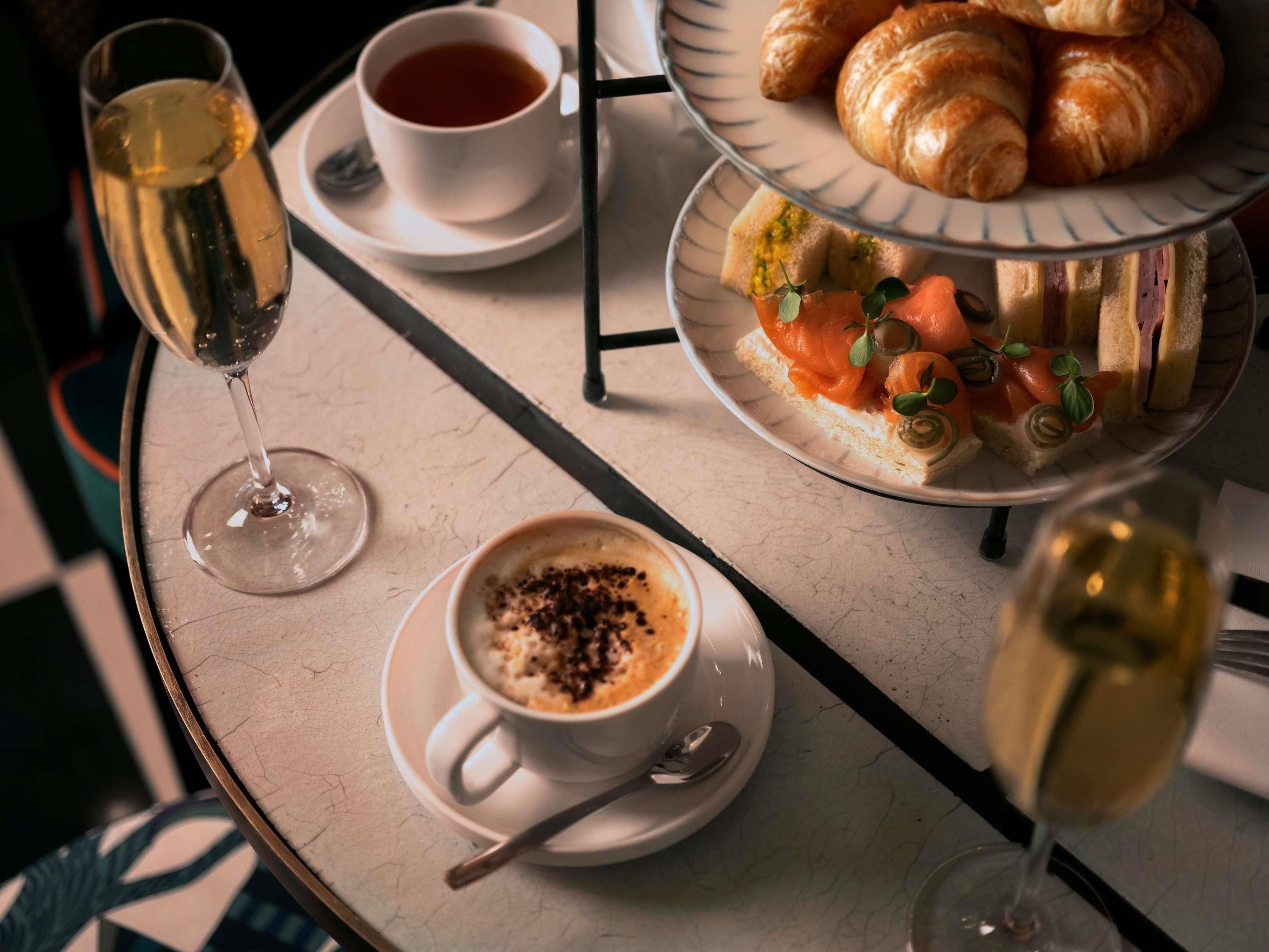 Una mesa con una copa de champán, una taza de té, un plato de croissants y una bandeja con pasteles y salmón ahumado.