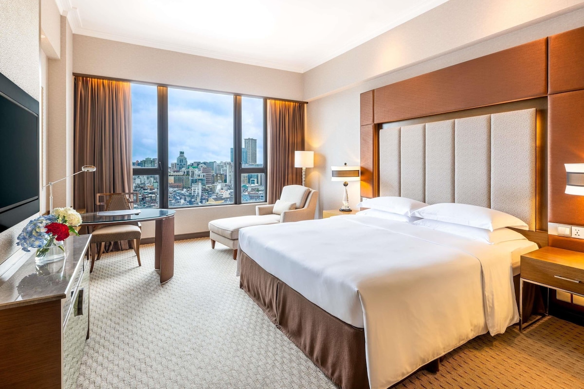 Una camera d'albergo con un letto grande, una scrivania con una sedia, una TV e vista sulla città.