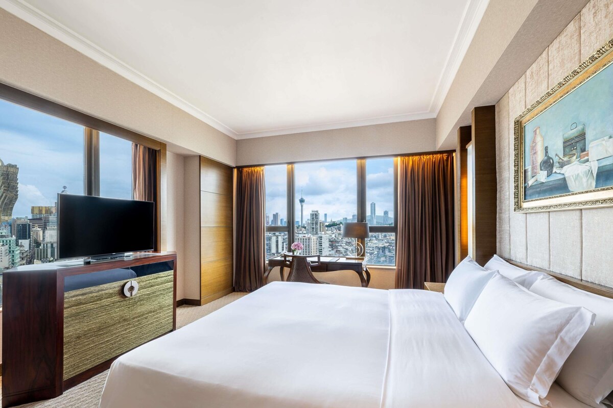 A hotel room with a large bed, a TV, a desk, and a view of the city skyline.