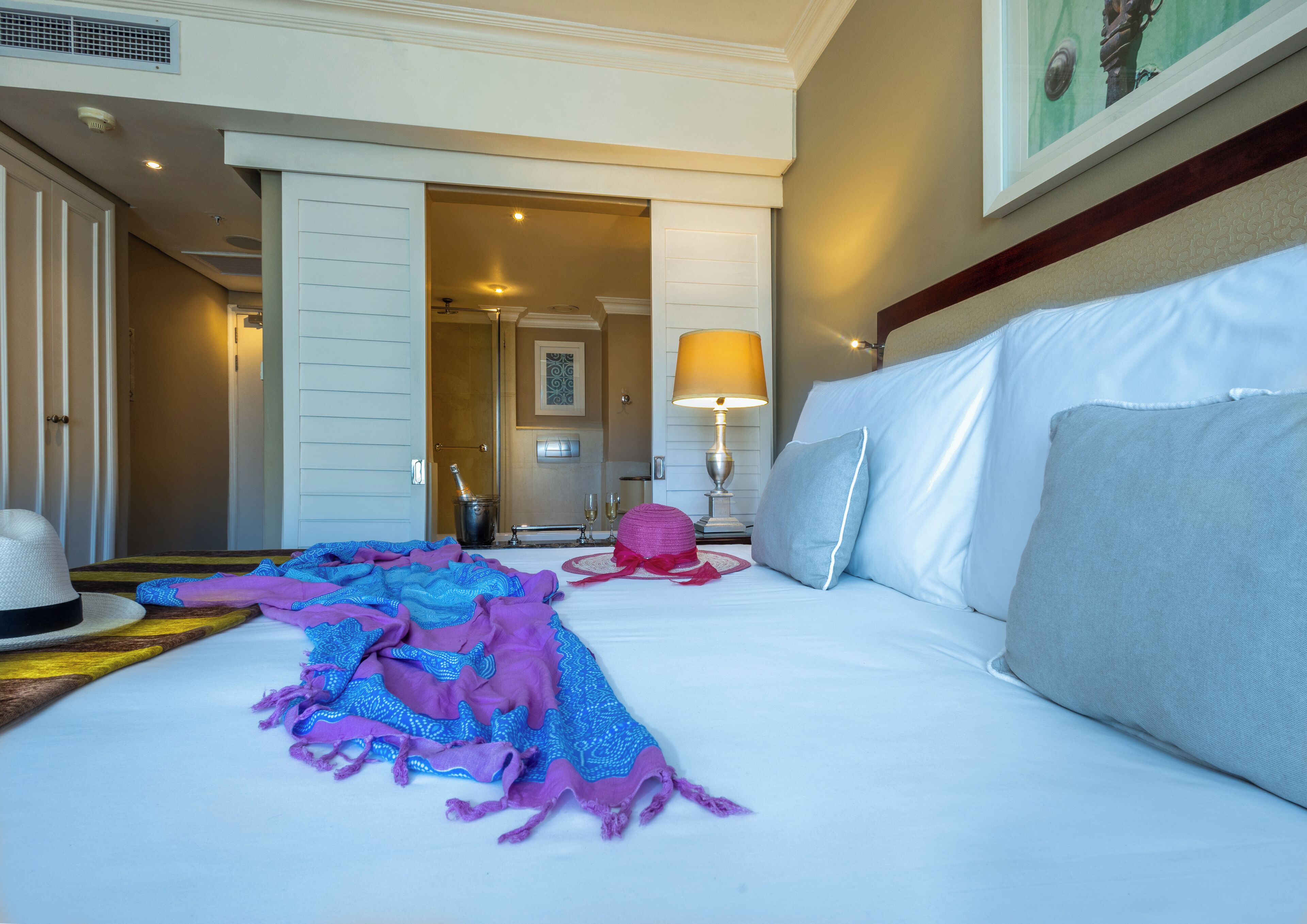 A hotel room with a bed, a nightstand, a lamp, and a door leading to another room.