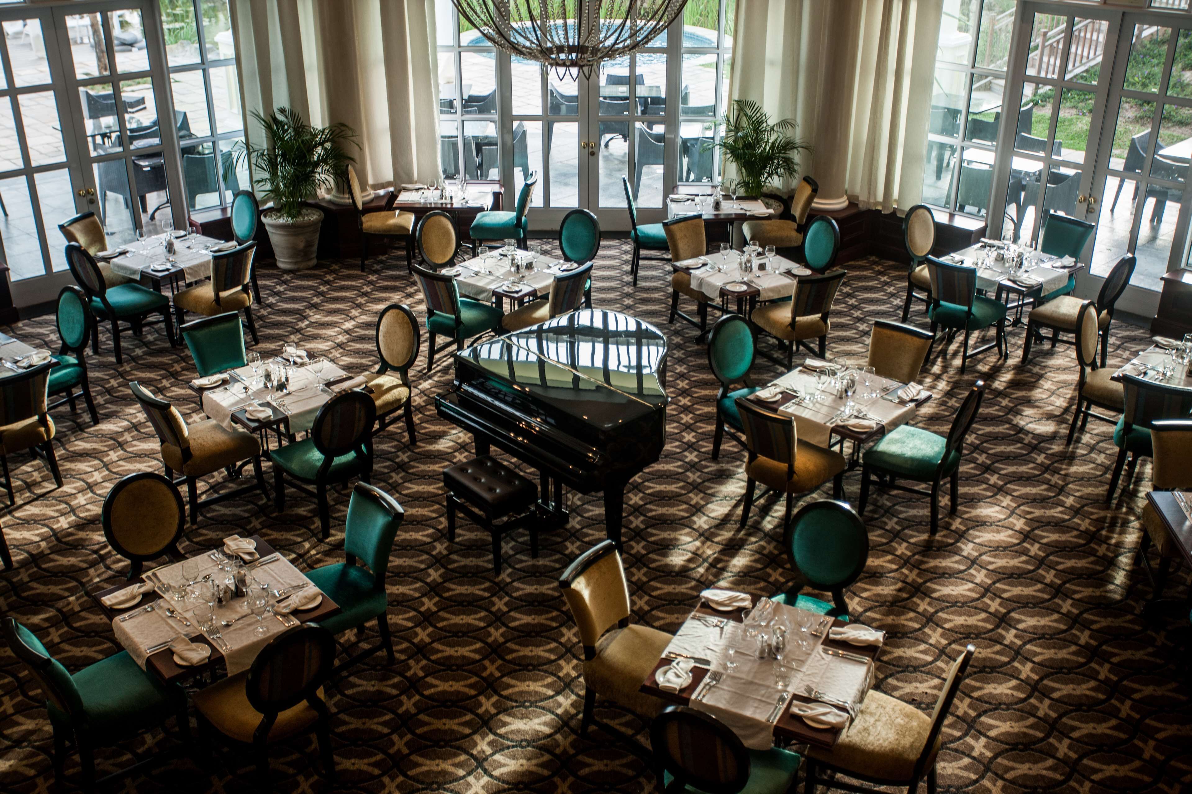 Een restaurant met een kamerbreed tapijt, groenbeige stoelen, tafels met witte tafelkleden en een vleugel in het midden.
