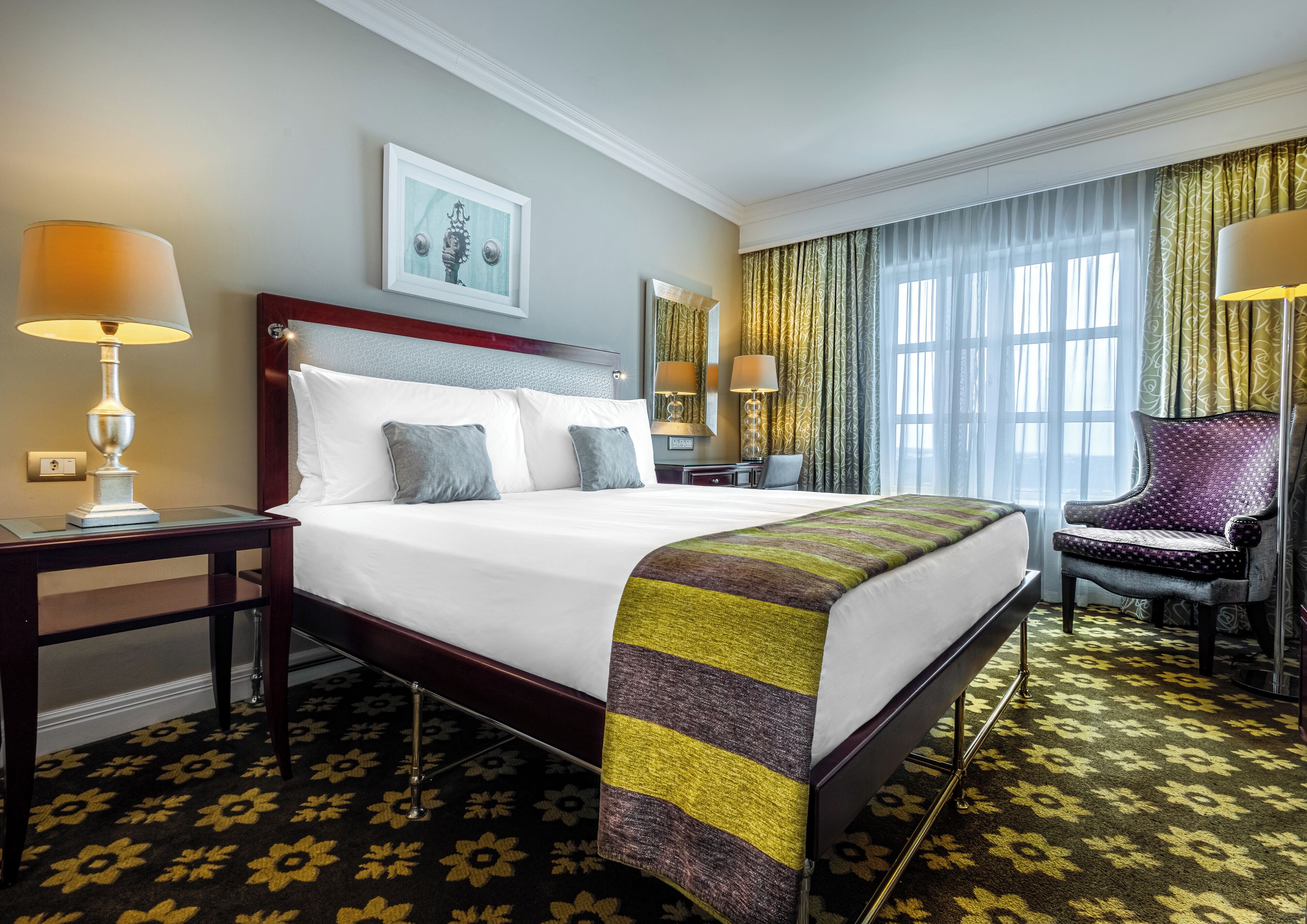 A hotel room with a large bed, two bedside lamps, a chair, a desk, and a window with curtains.