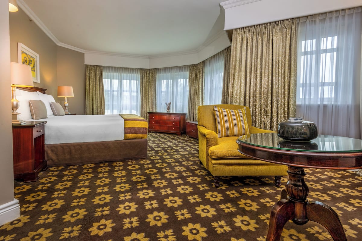 A hotel room with a large bed, a yellow armchair, a glass table, and a patterned carpet.