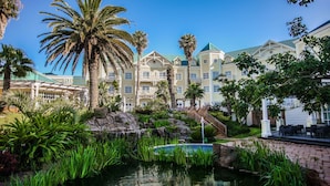 A resort with a pool surrounded by tropical plants and palm trees, a stone bridge, and a building with a green roof. - Hemingways Hotel  (East London)