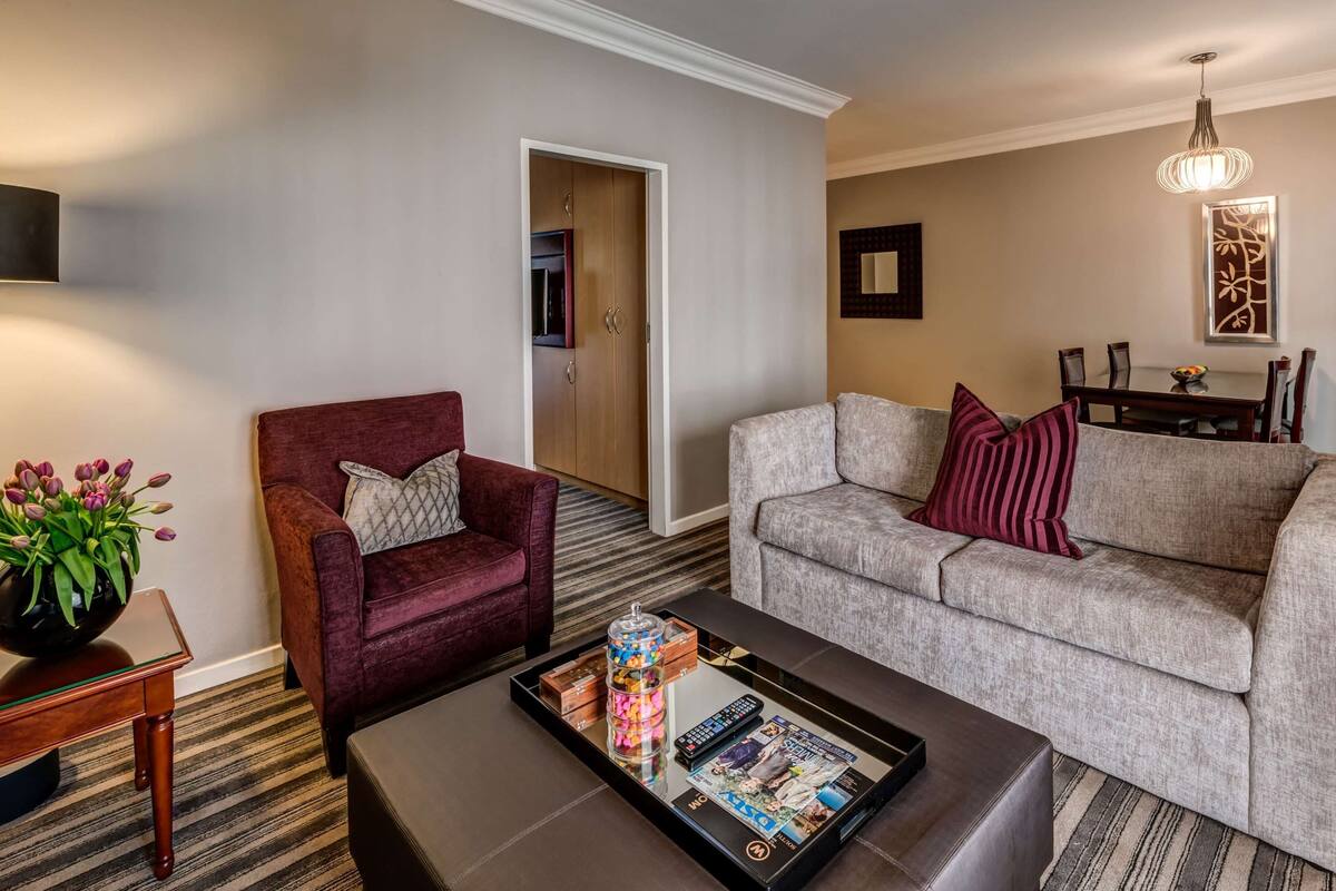Ein Hotelzimmer mit einem roten Sessel, einem grauen Sofa, einem Couchtisch mit Zeitschriften und einer Ablage sowie einer Stehlampe und einem Essbereich mit einem Kronleuchter.