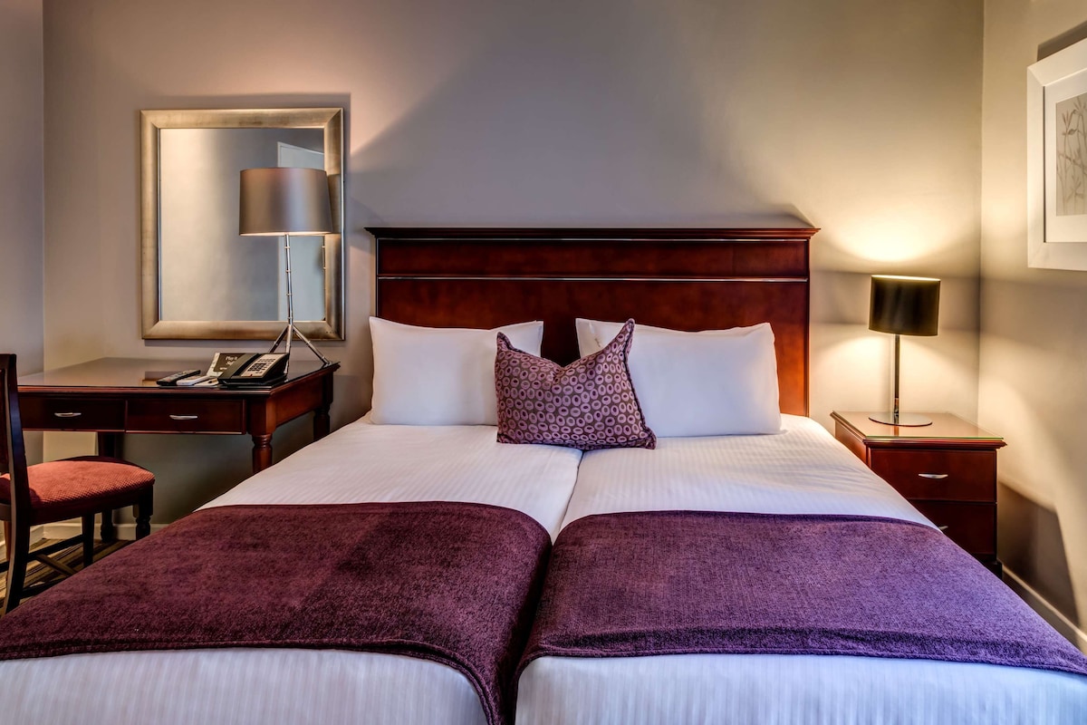 A hotel room with a double bed, two bedside tables, a desk with a chair, a mirror, and a framed picture on the wall.