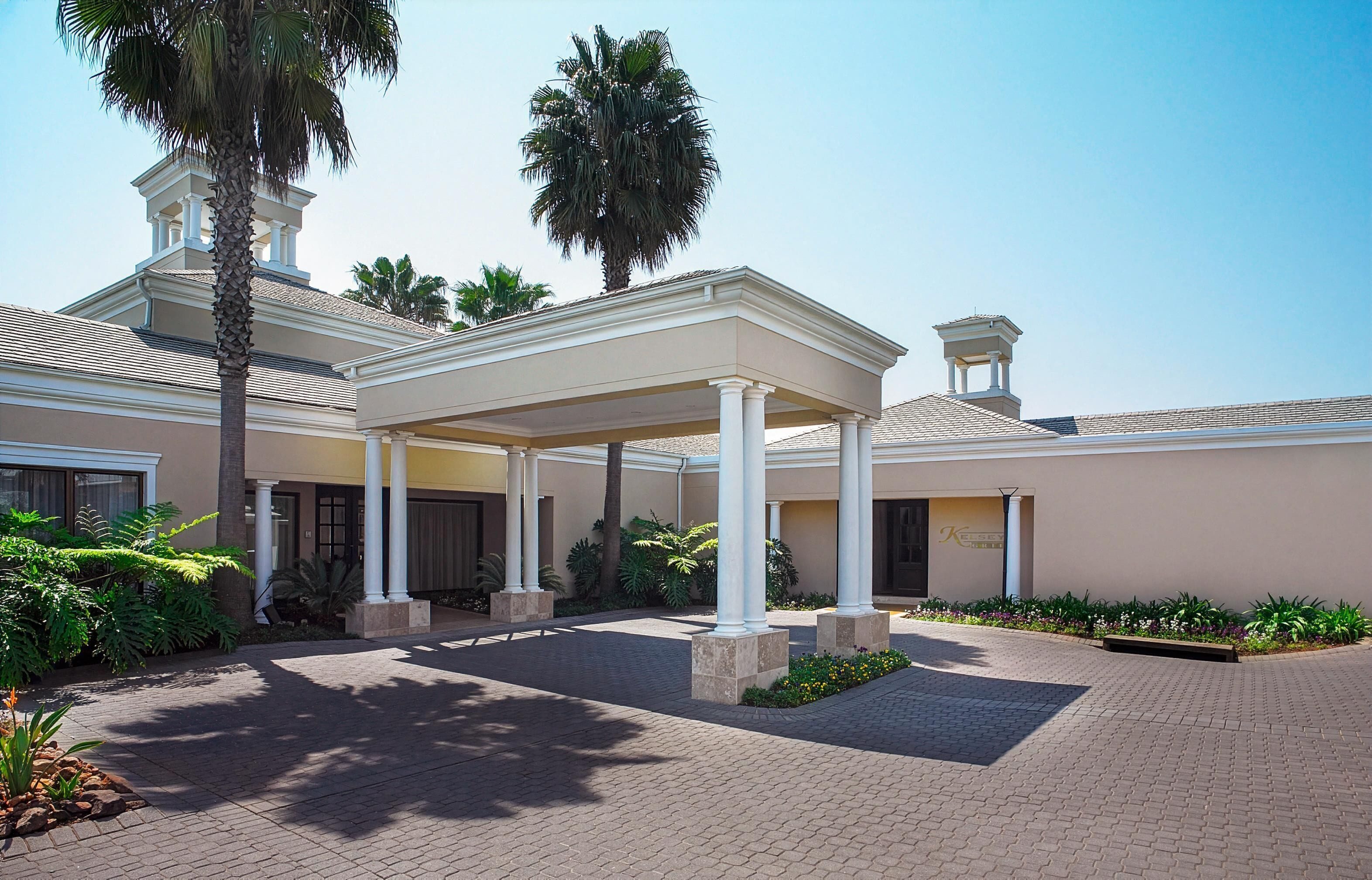A covered outdoor area with a central column structure, palm trees, and a building with a beige facade and a tiled roof.
