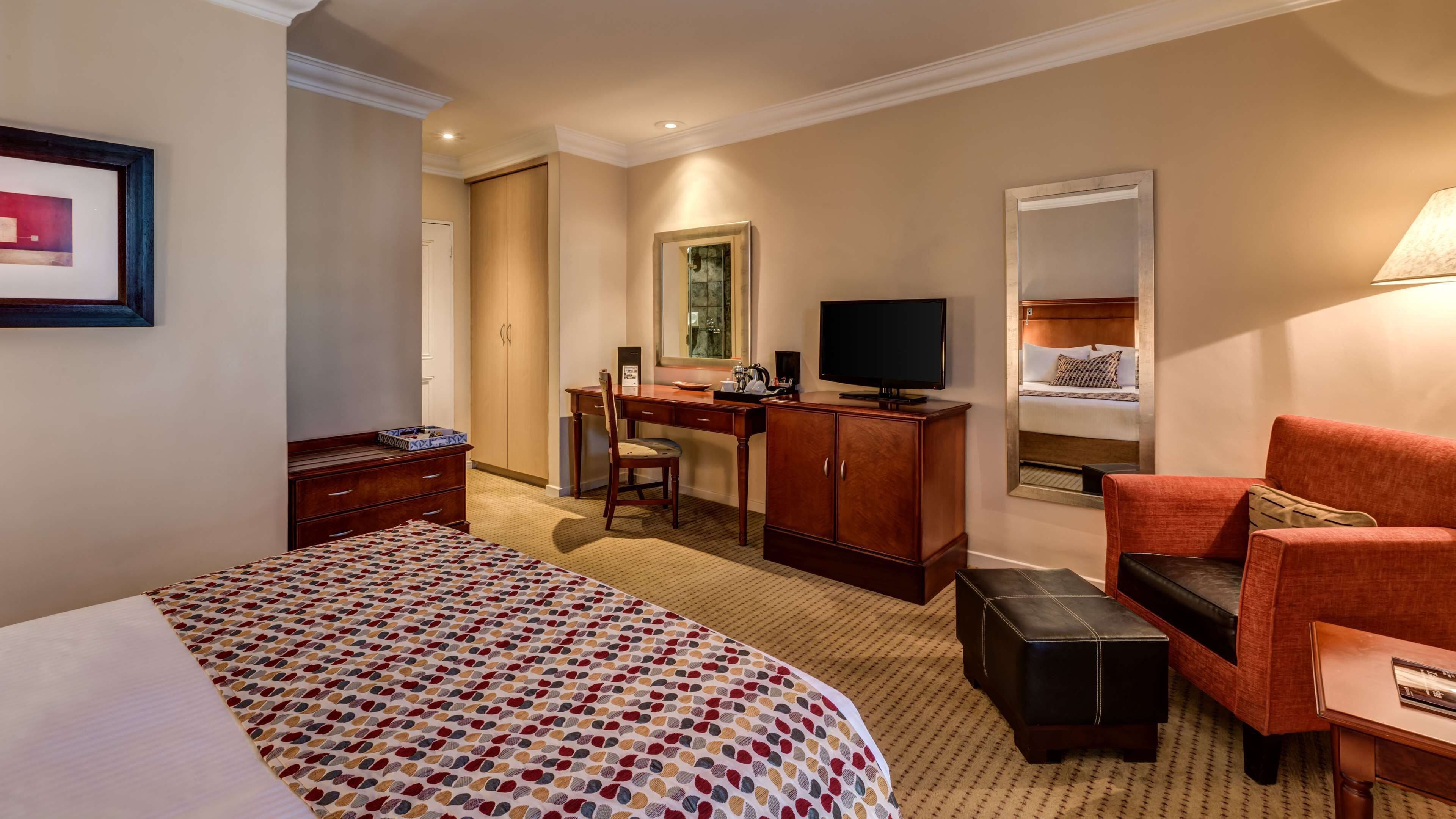 Een hotelkamer met een groot bed, een bureau met stoel, een televisie, een spiegel en een rode fauteuil.