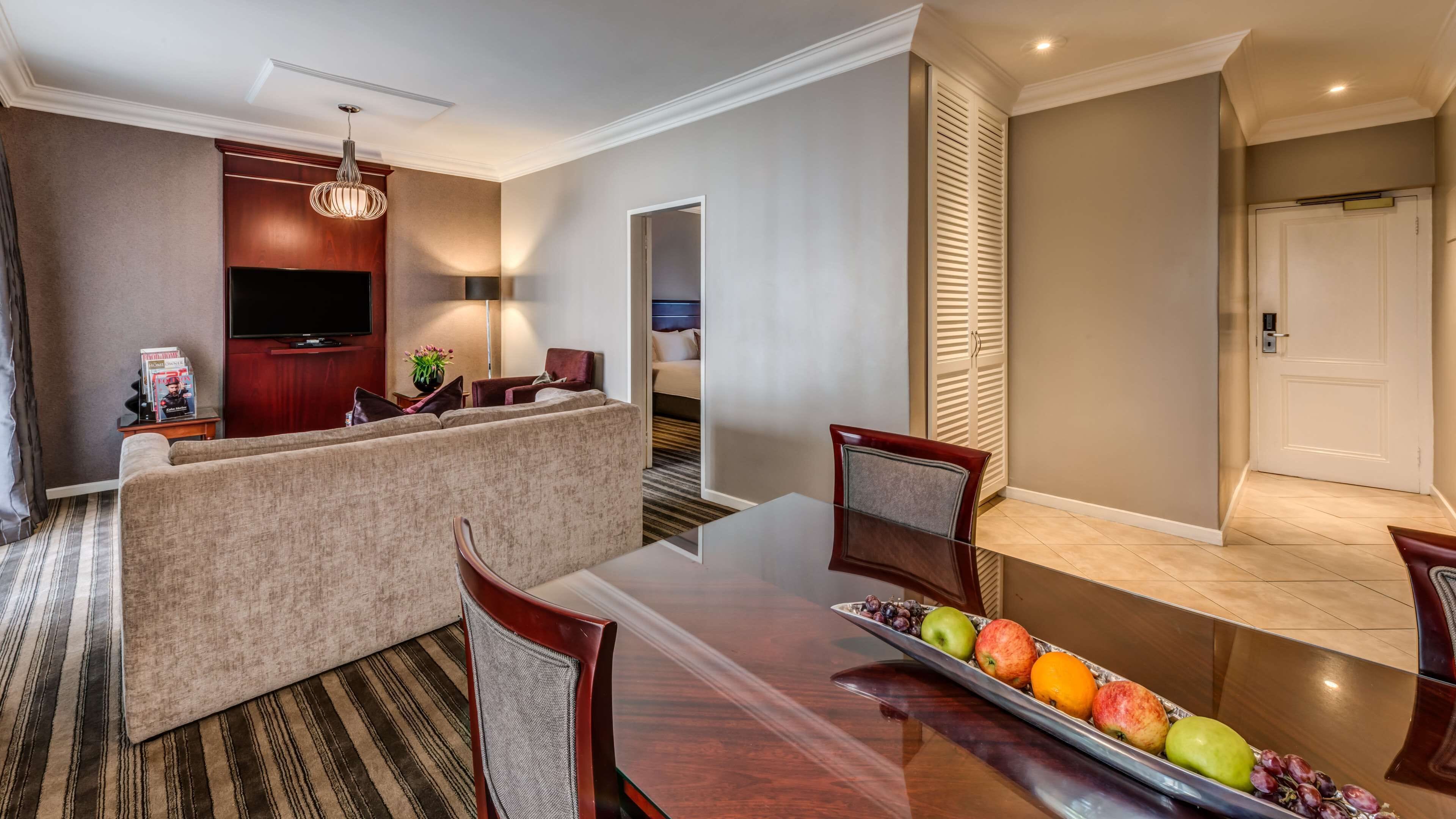 A hotel room with a dining area featuring a long table, chairs, and a fruit tray. There is a sofa and a TV in the living area.