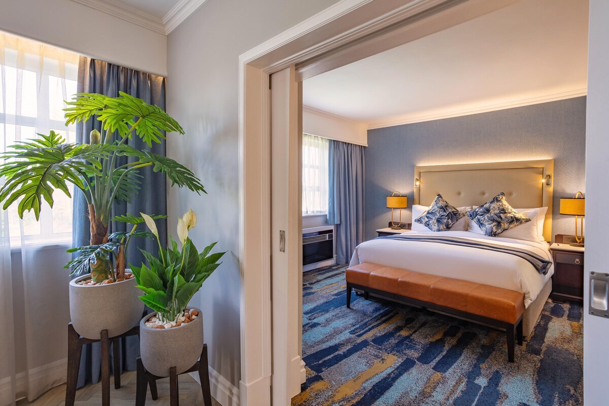A hotel room with a large bed, a nightstand, two bedside lamps, a headboard, and a potted plant.