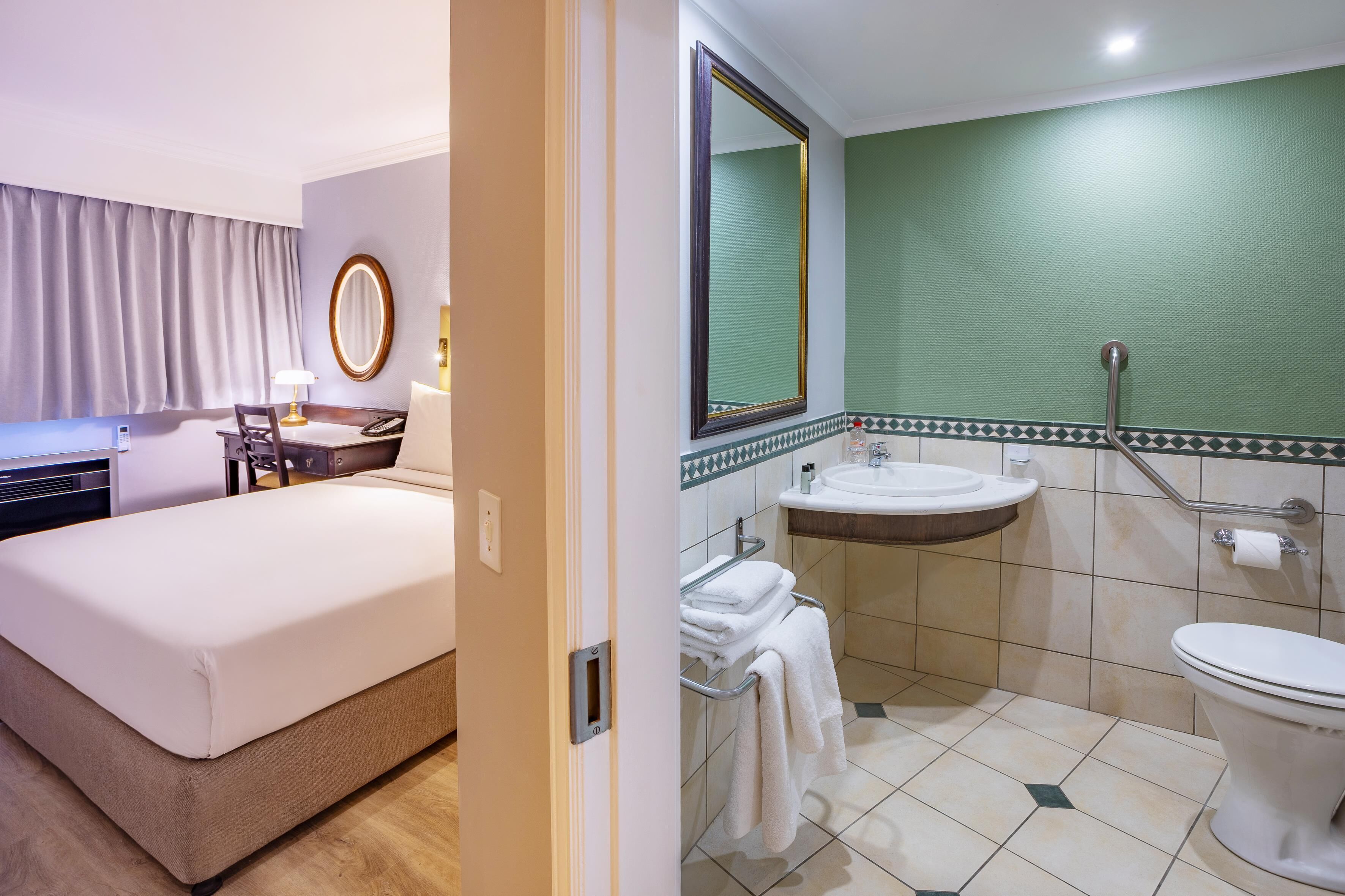 Habitación de hotel con cama, escritorio con lámpara, baño con lavamanos, espejo y retrete.