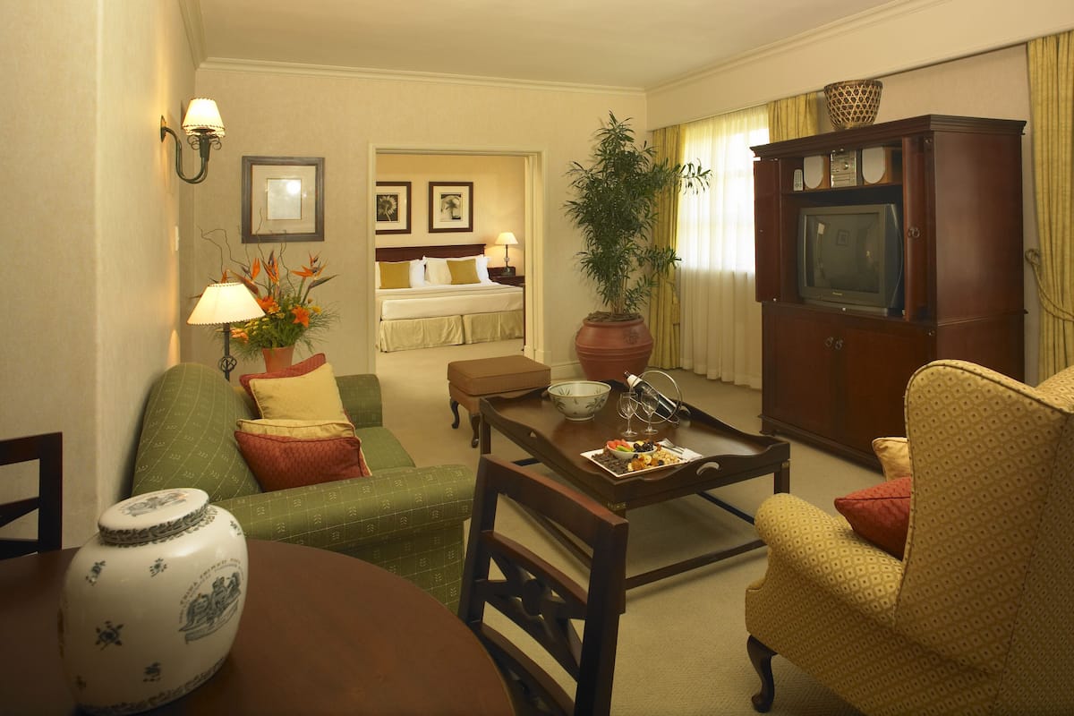 A hotel room with a green sofa, a yellow armchair, a wooden coffee table, a TV cabinet, and a dining table with a vase.