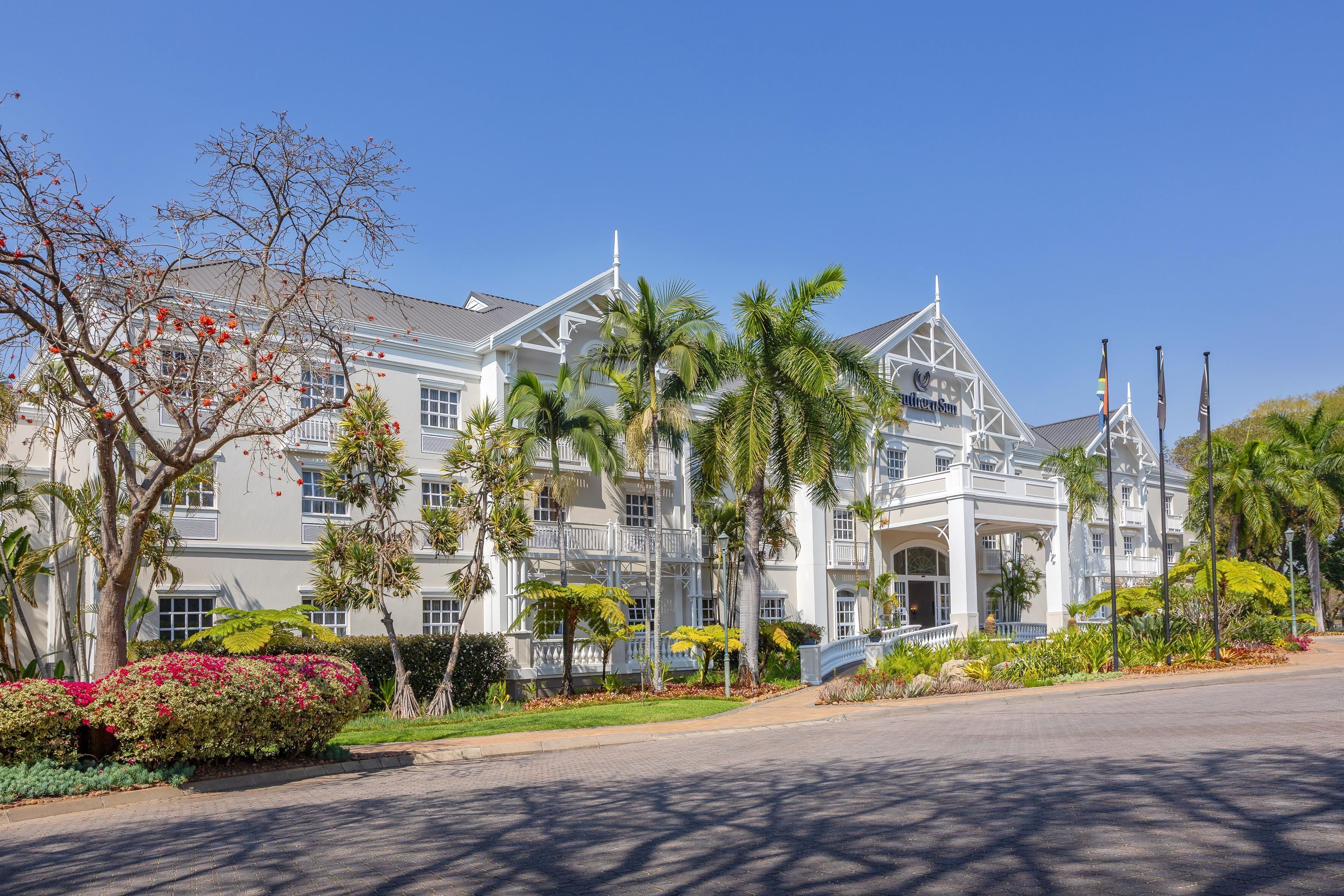 Um hotel grande e branco, com vários andares, arquitetura clássica, com varandas e um jardim bem cuidado com plantas tropicais.
