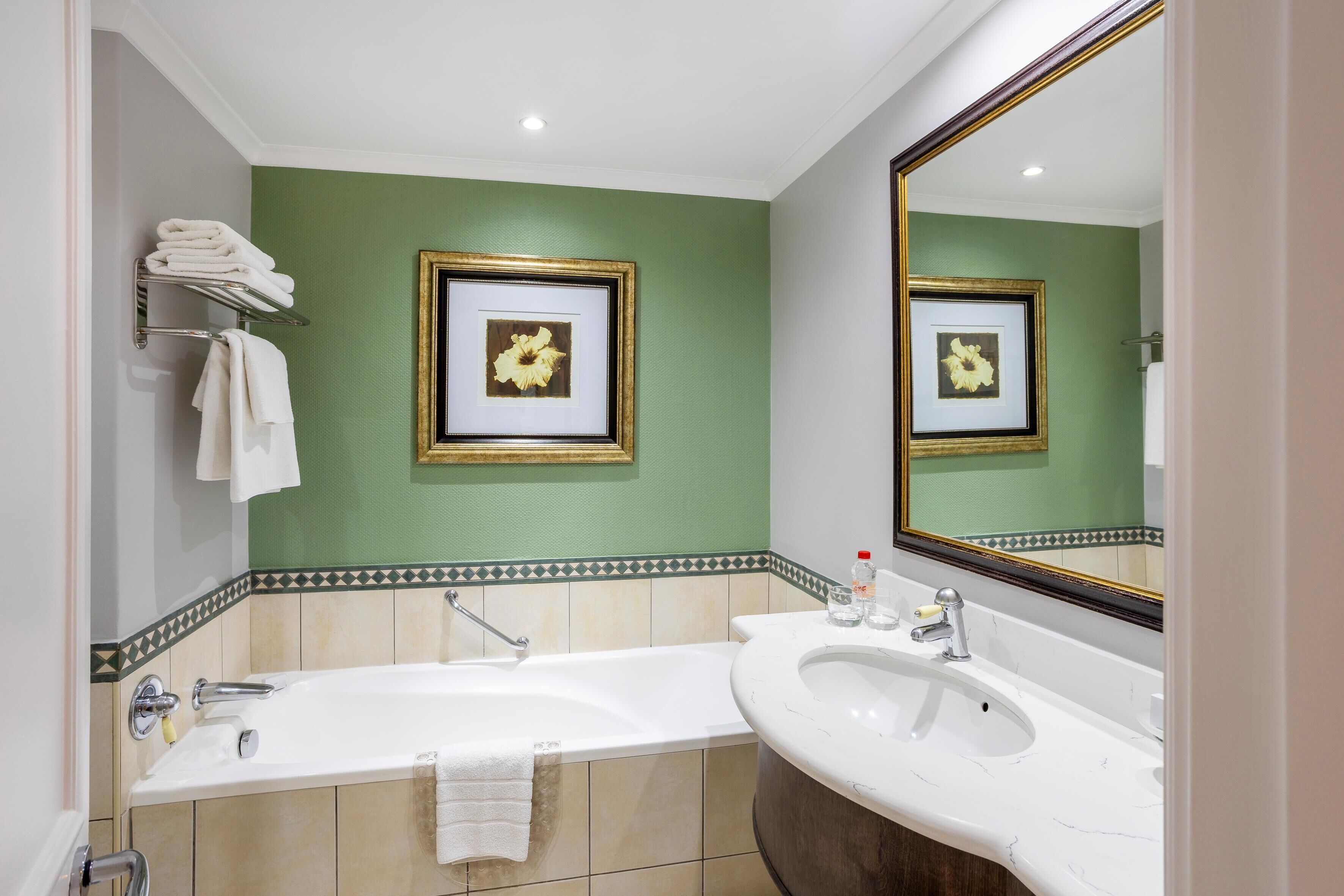 Un baño con una bañera grande, un lavamanos y un espejo. Hay toallas en un perchero y un cuadro en la pared.