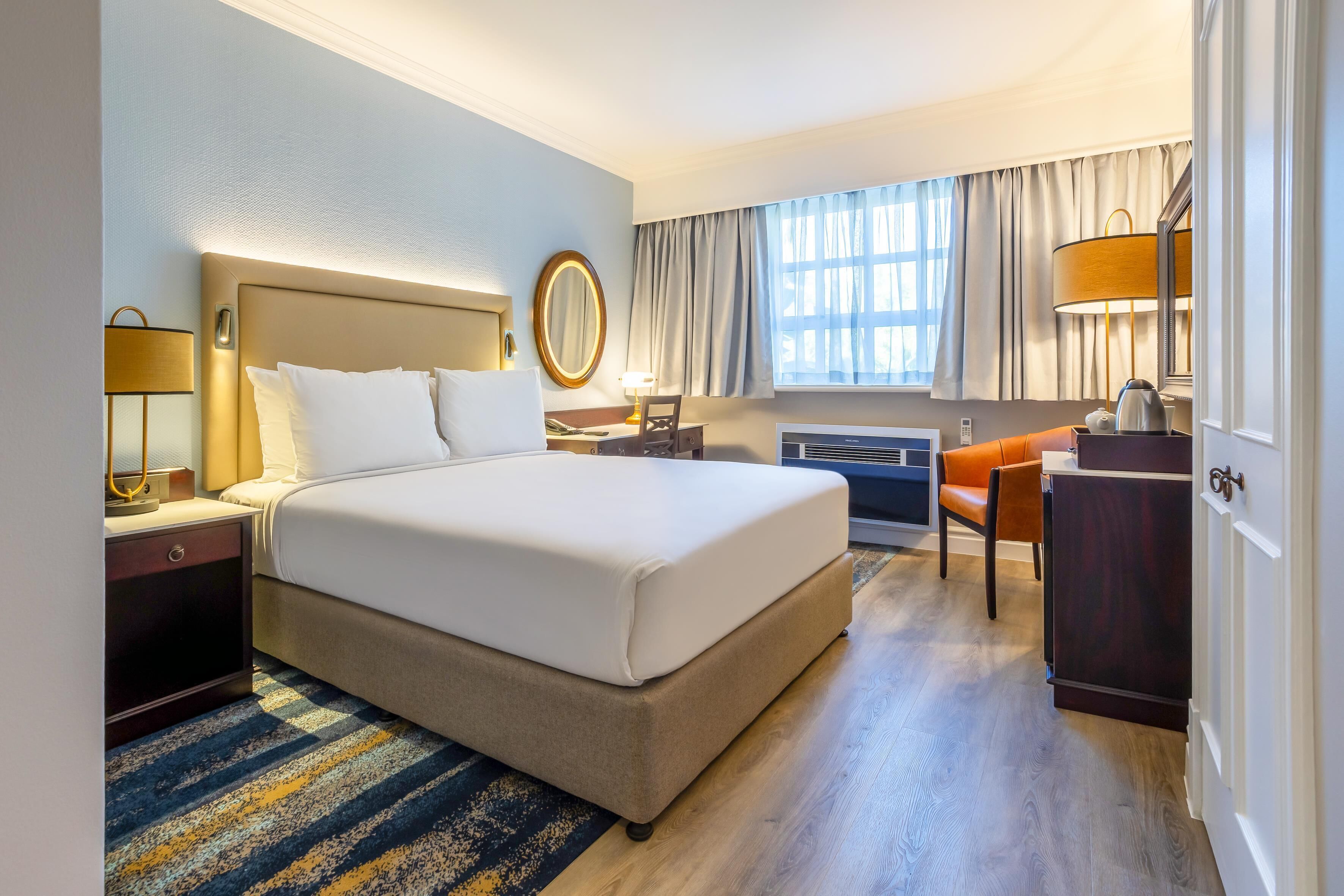A hotel room with a large bed, a desk, a chair, a lamp, a mirror, and a window with curtains.