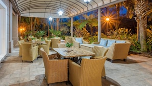 An outdoor patio with wicker furniture and a stone floor, surrounded by tropical plants and palm trees. - Southern Sun Mbombela (Mbombela)