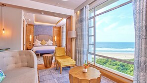 Habitación de hotel con cama, sofá, sillón, una mesa de centro redonda y un ventanal grande con vista a la playa.